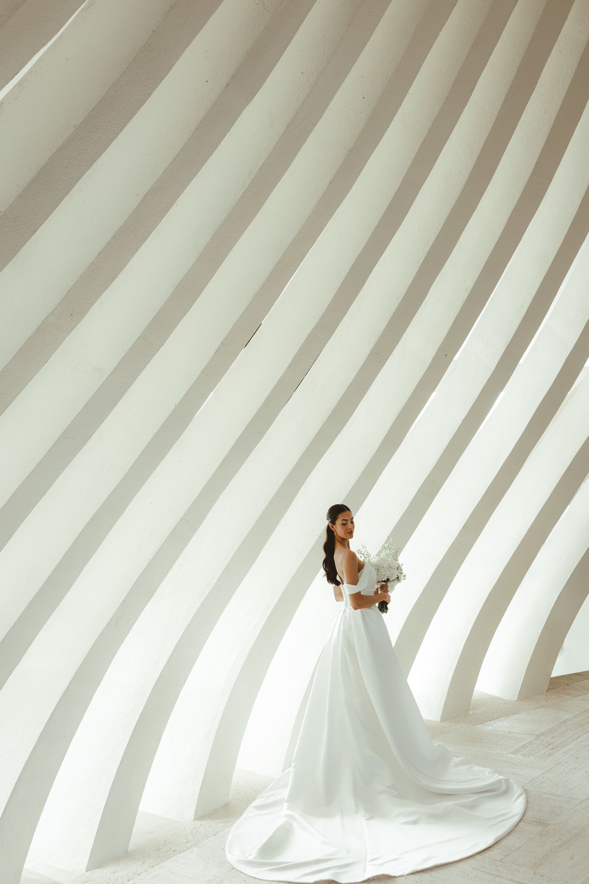 A woman in a white wedding gown holding a bouquet of white flowers stands against a modern, curved architectural background with diagonal white slats.