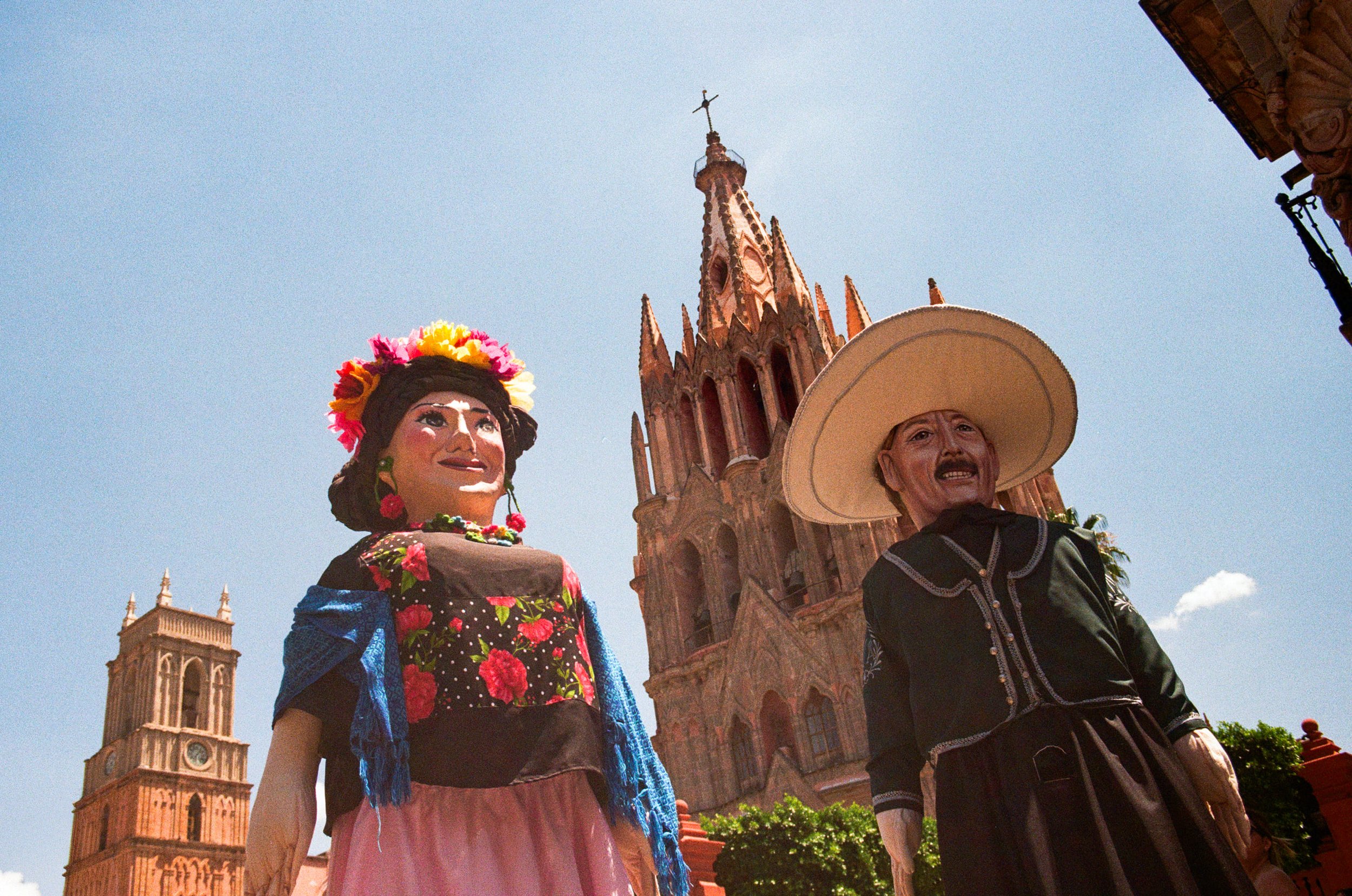Colorful large figurines of a woman and a man dressed in traditional Mexican clothing are in front of San Miguel de Allende Church, set against a blue sky during a wedding celebration.