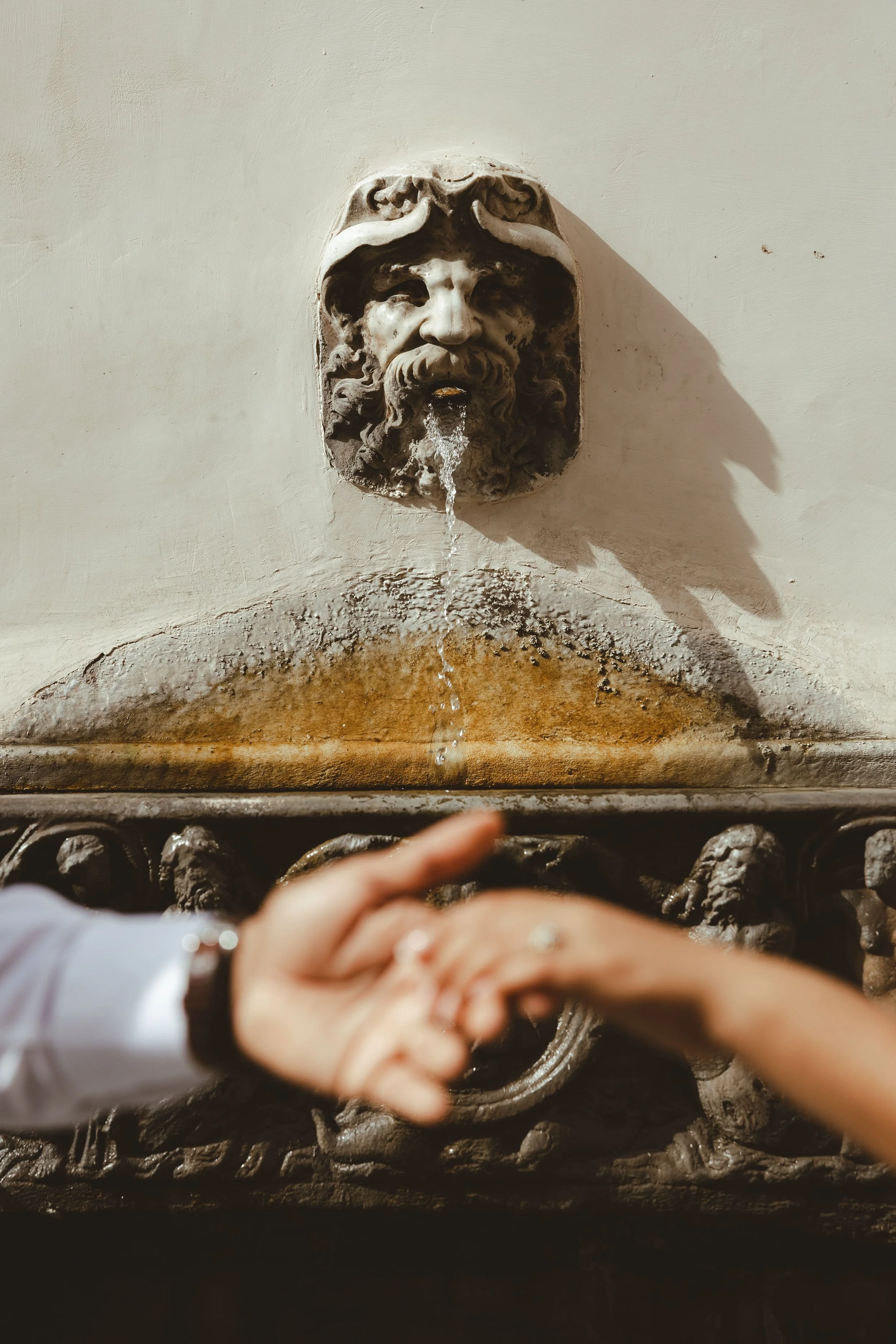A decorative wall fountain with a face sculpture from which water flows, featuring a beard and a mustache, mounted on a white wall with a shadow cast to the right.