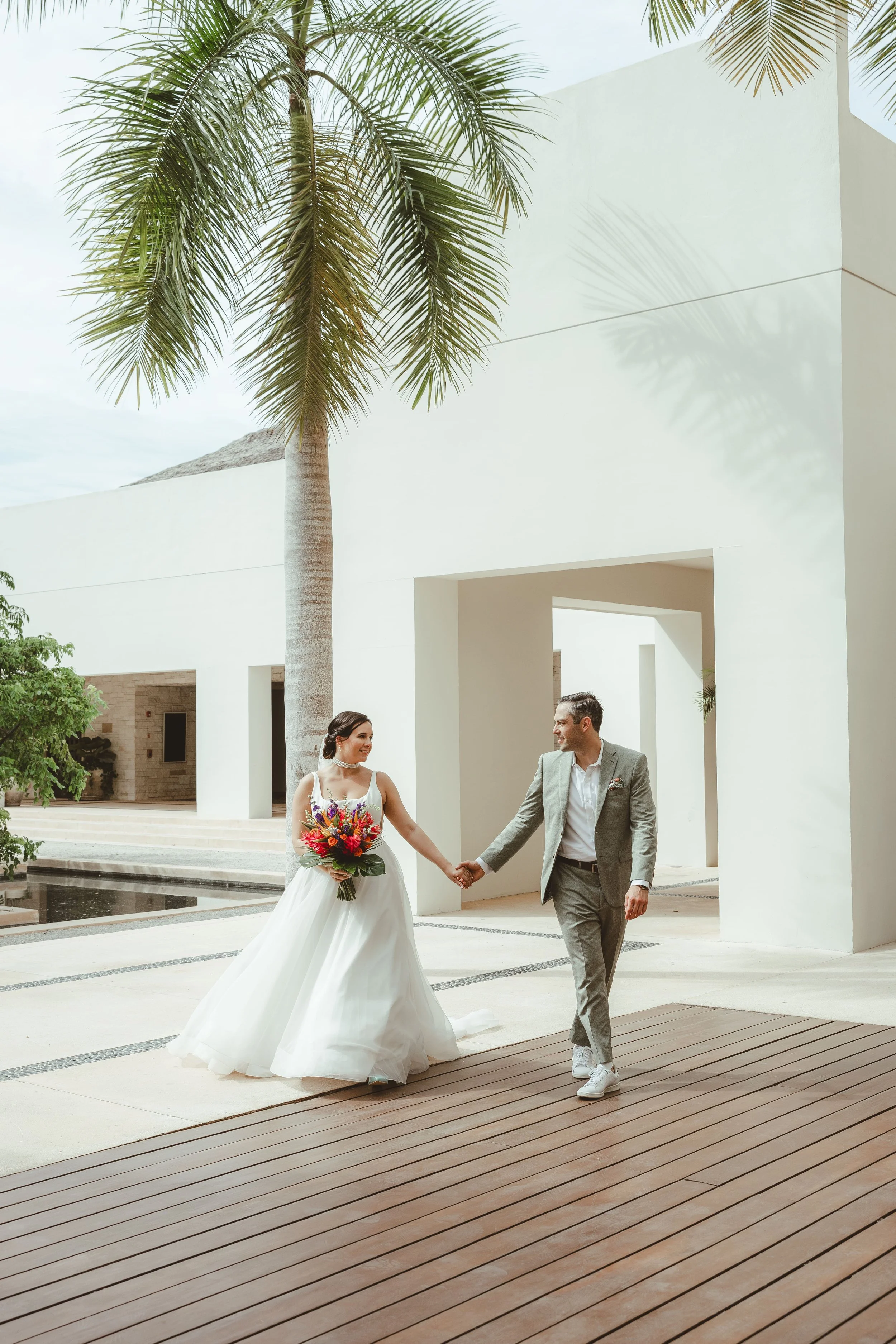 A bride and groom walking hand in hand outdoors during a wedding, with a modern white building, palm trees, and water feature in the background.