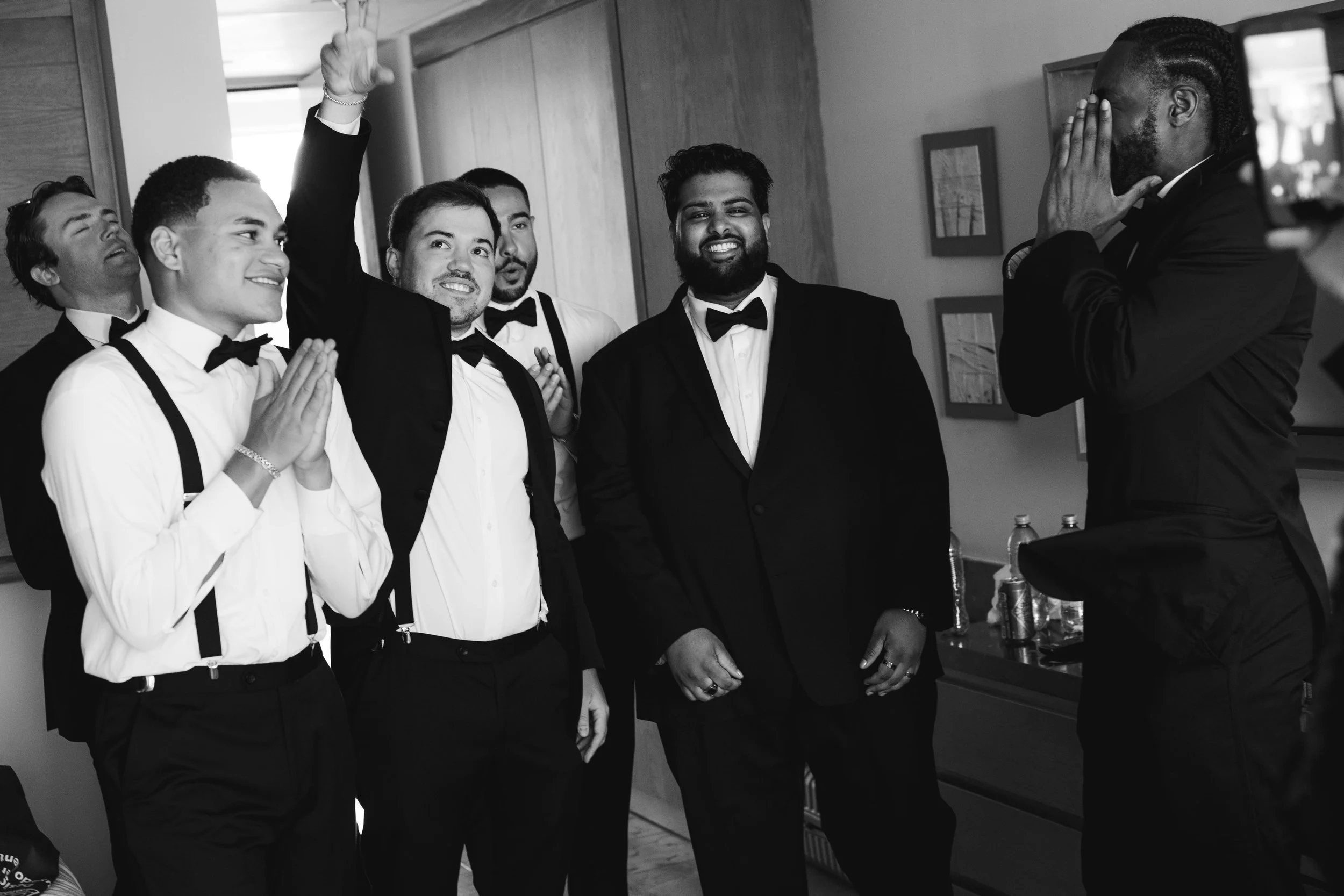 Group of men dressed in formal tuxedos, some with suspenders and bow ties, gathered in a room, smiling and posing for a photo. One man covers his face with his hands, another raises his arm making a peace sign, and others are smiling or clapping.