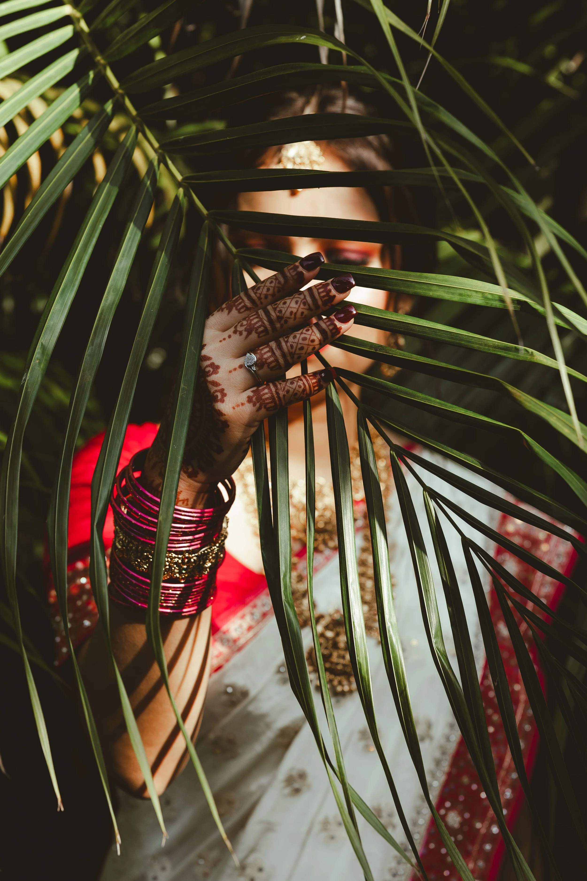 A woman wearing traditional Indian attire and henna on her hands, partially hidden behind green palm leaves, with decorative jewelry and a vibrant red sari.