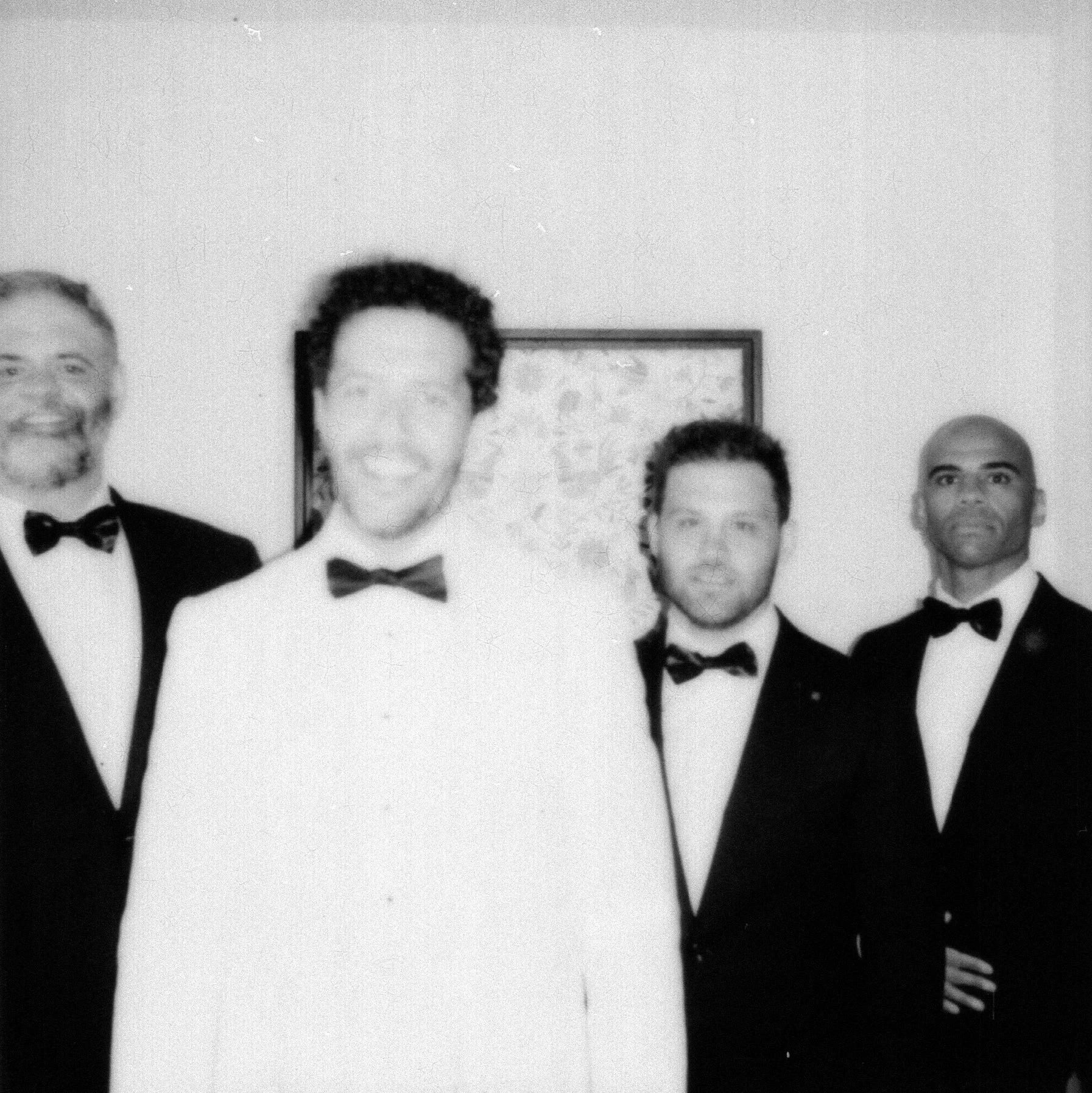 Black and white photo of a groom and his groomsmen dressed in tuxedos and bow ties, standing side by side against a plain background with a map or framed picture behind them.