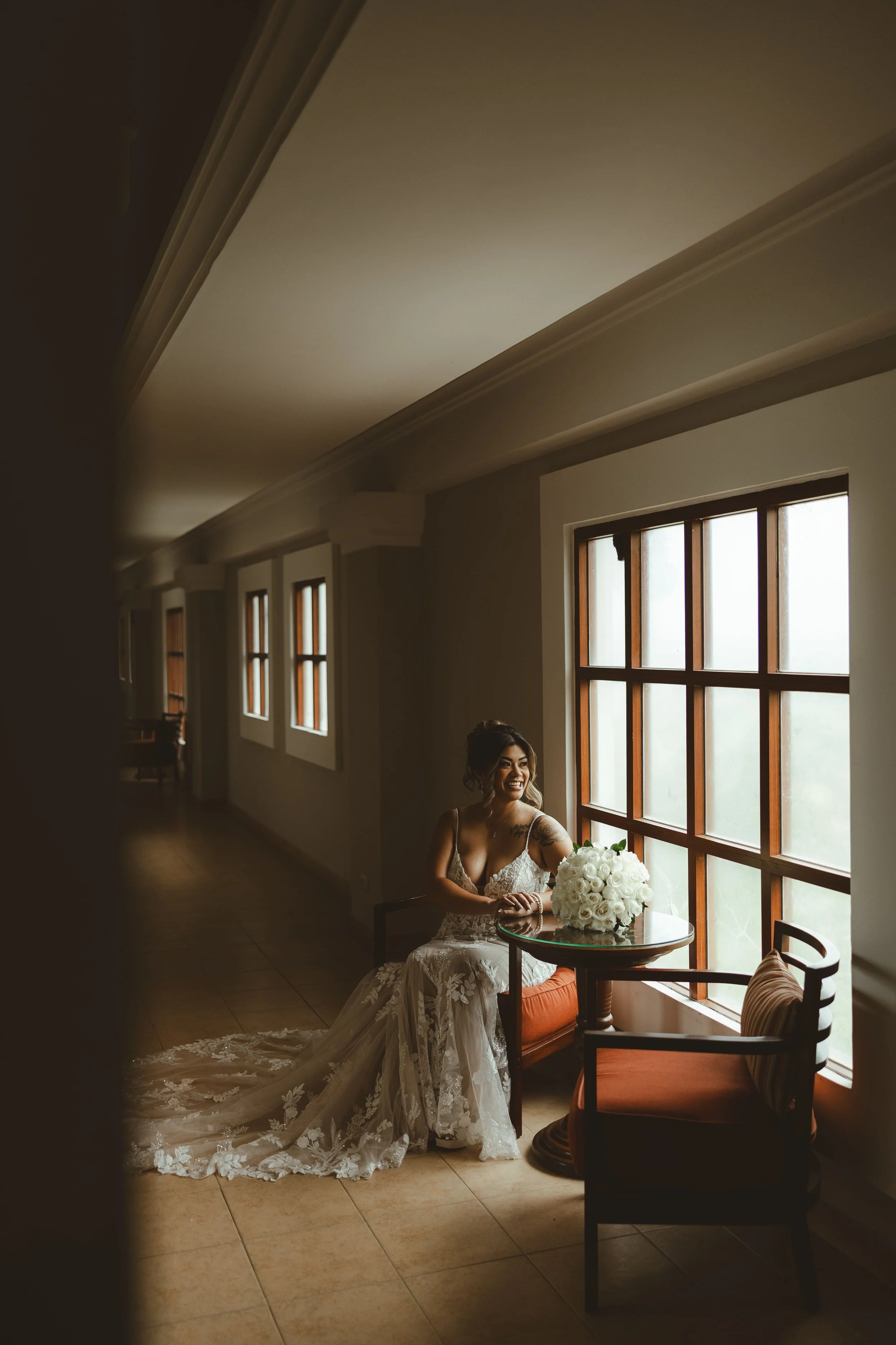 A bride with dark hair in a wedding dress sitting by a large window with natural light, holding a bouquet of white roses, smiling in a warmly lit room with wooden framed windows and terracotta tile flooring.