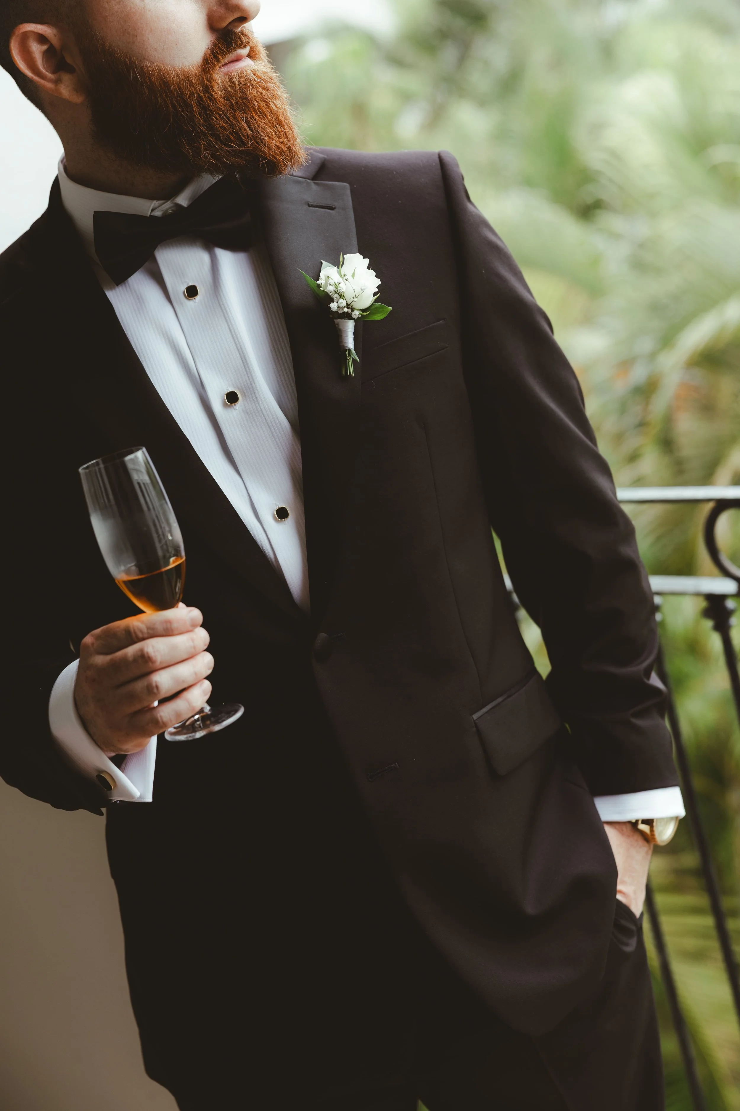 A man in a black tuxedo with a white shirt and black bow tie, holding a glass of champagne. He has a red beard and a boutonnière with white flowers on his lapel. The background is blurred greenery.