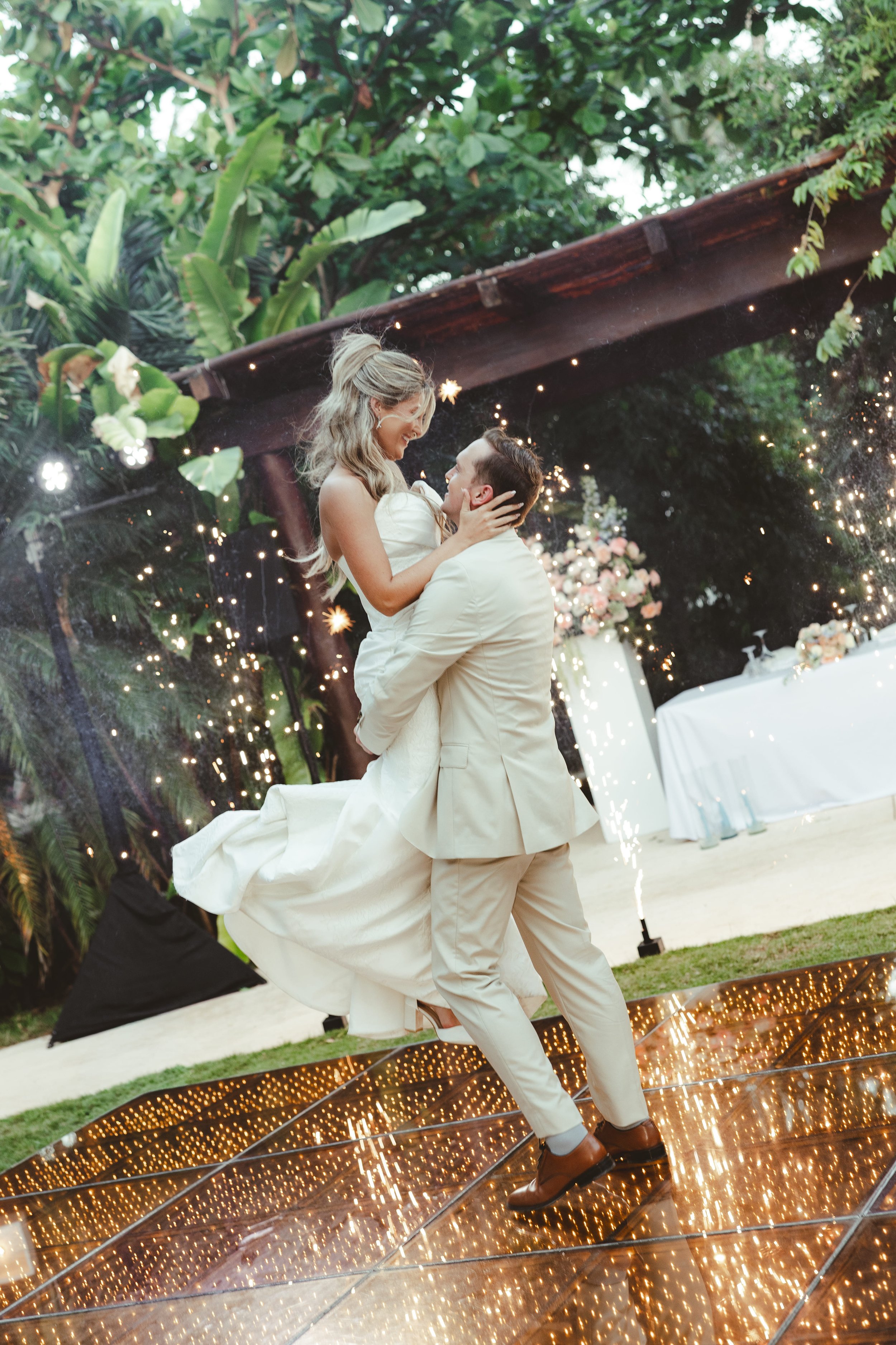 A couple at a wedding dance outdoors under string lights, with lush green trees in the background, and sparks from a fountain or firework display near the ground.
