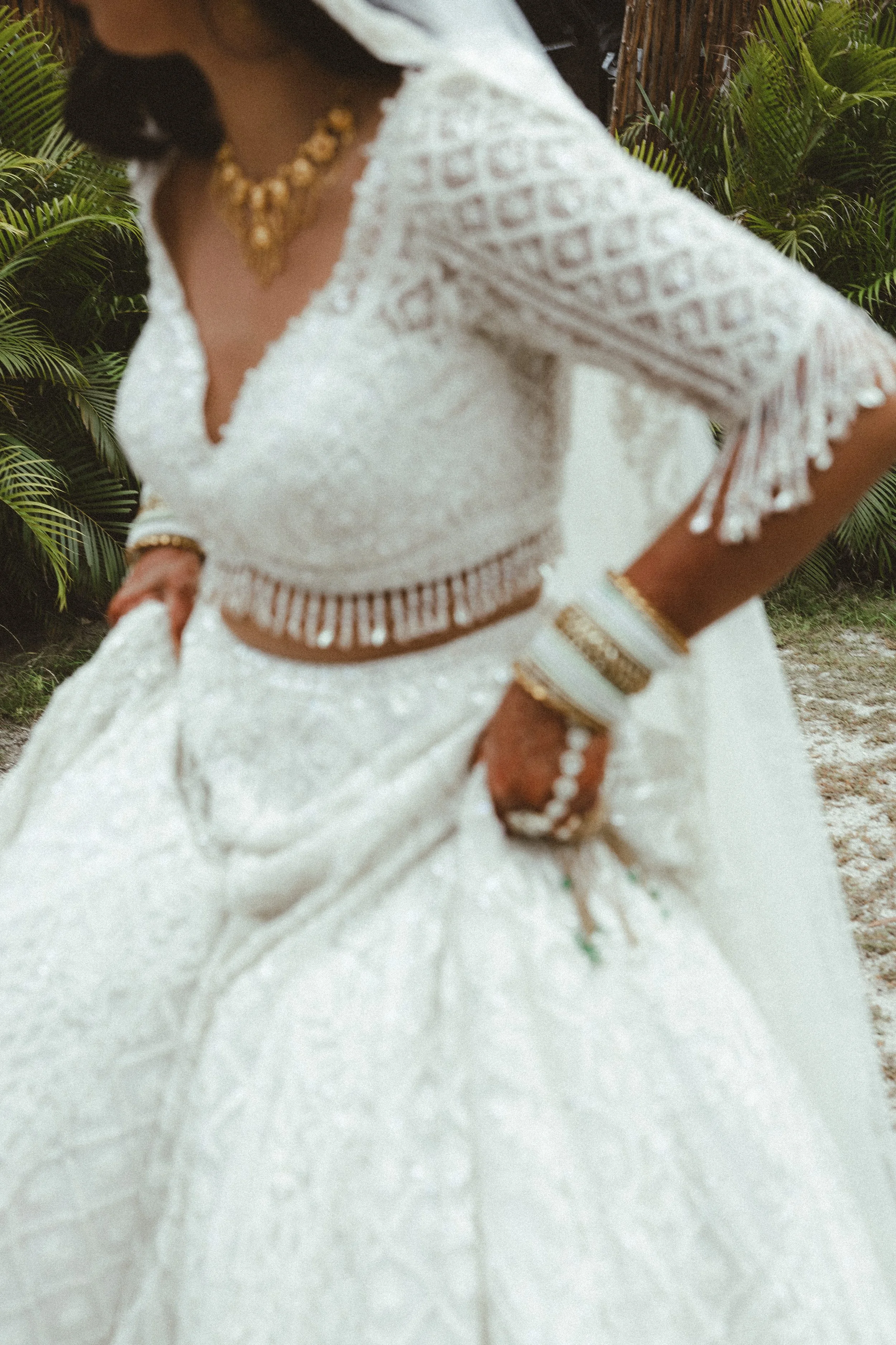 A bride wearing a white lace dress with gold jewelry, including a necklace, bracelet, and bangles, standing outdoors with green foliage in the background. It's a south asian wedding attire.