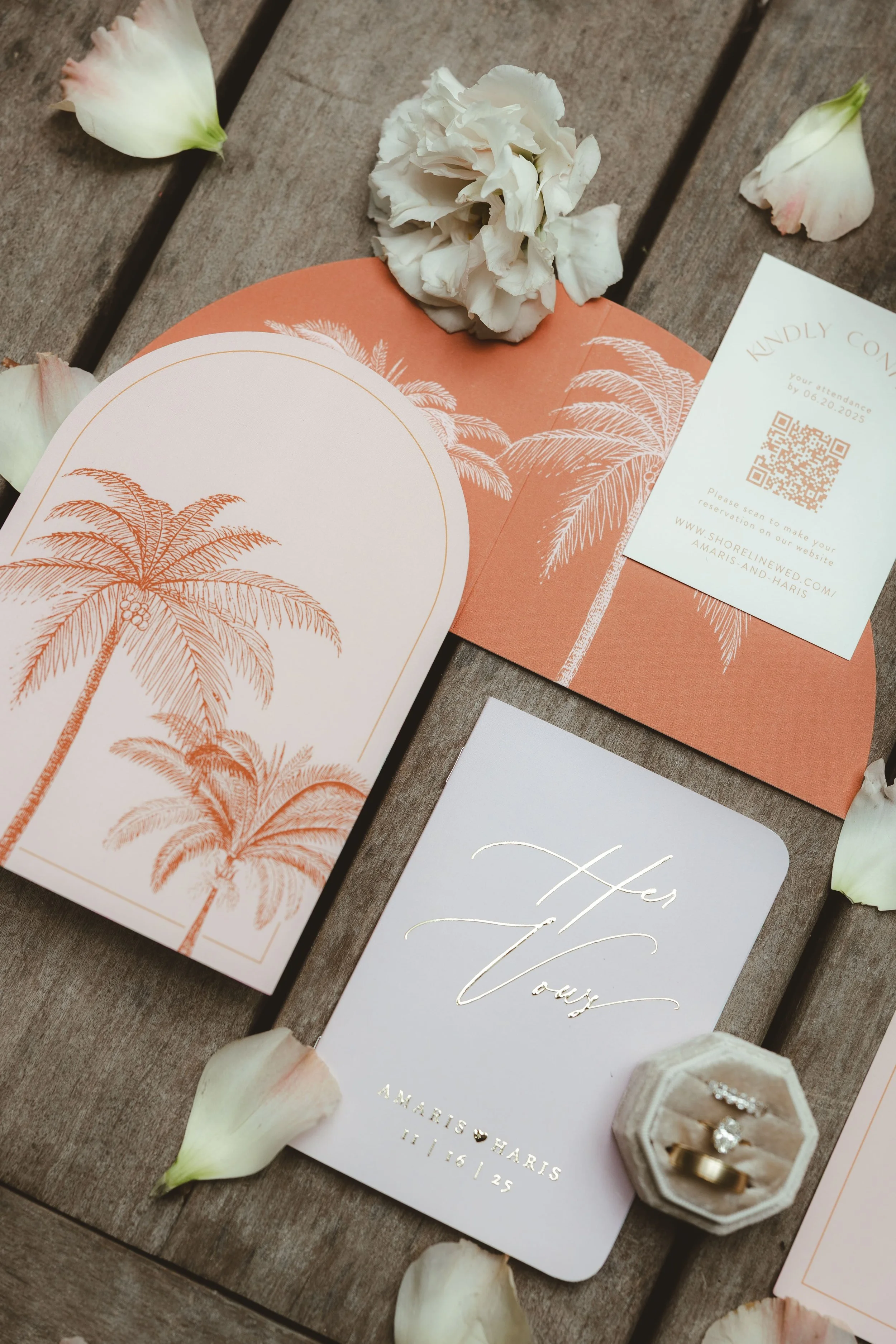 A wedding invitation suite and a small velvet ring box containing wedding rings, arranged on a wooden surface with scattered white flower petals. Details of a wedding.