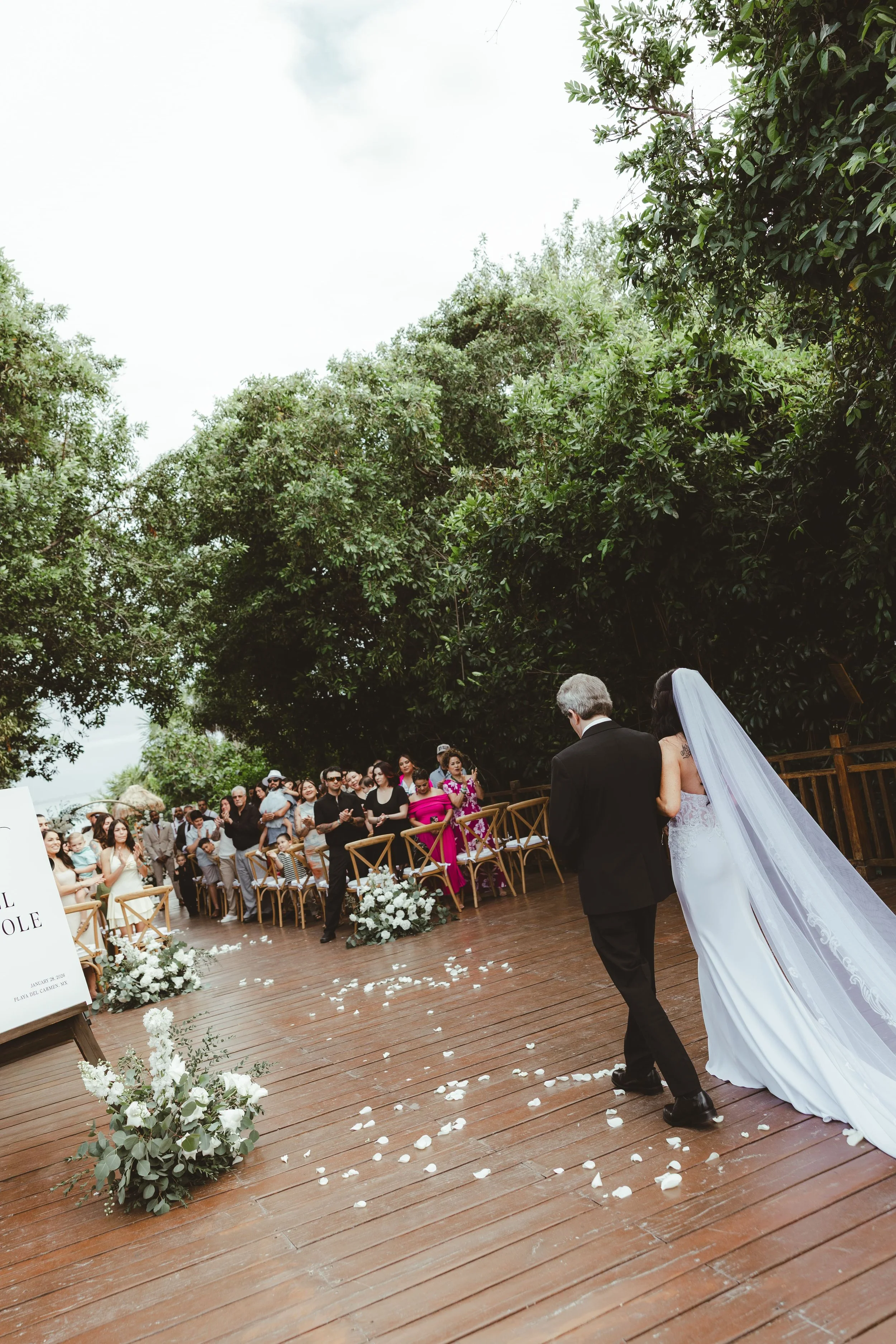 A bride and groom walking down the aisle at their outdoor wedding ceremony, with guests seated on either side and white flower decorations on the wooden floor. Green trees surround the scene.