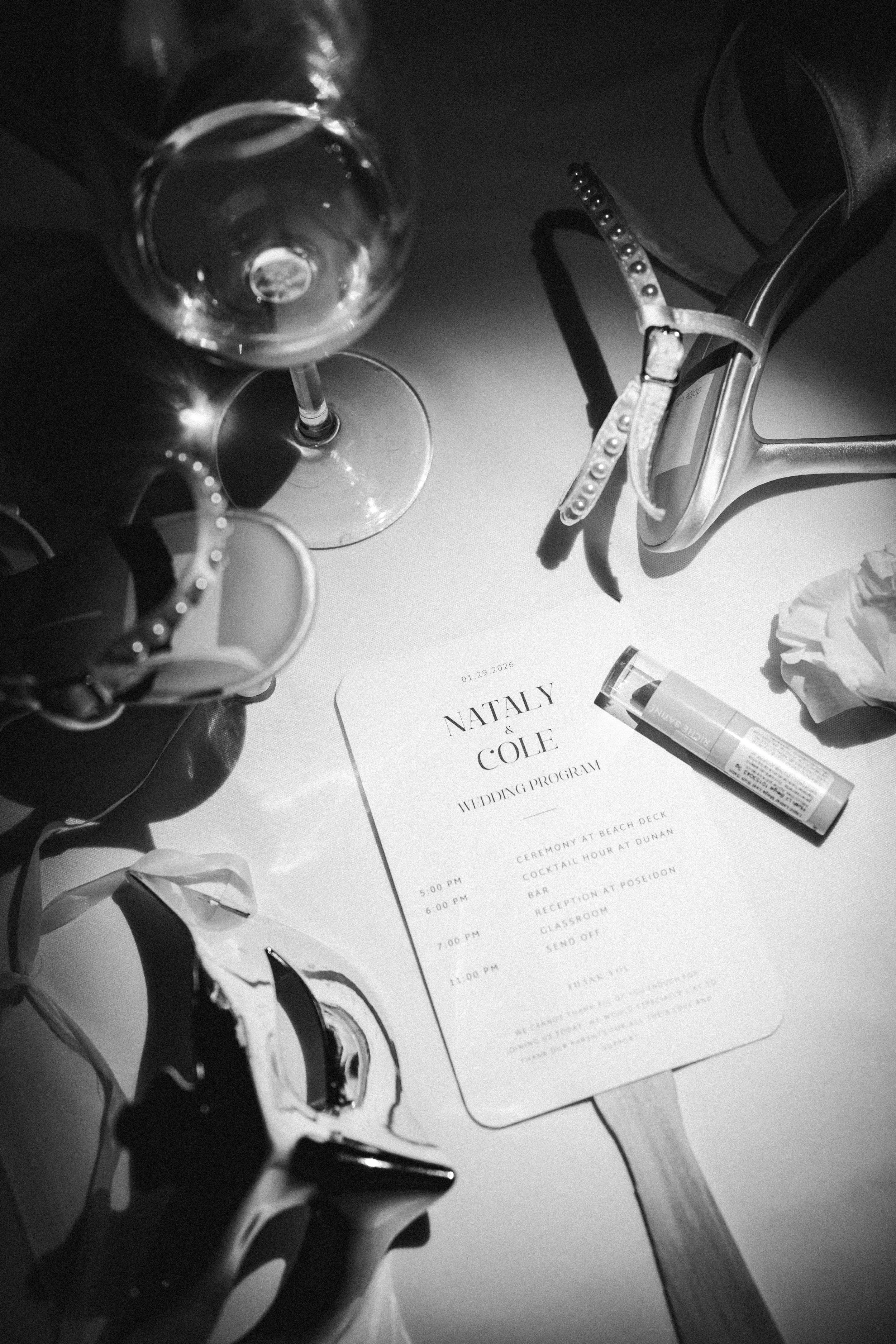 Photo of a table with a wedding program, a glass of water, a perfume bottle, and a pair of sunglasses. The wedding program is for Nataly and Cole, with a schedule of events. The sunglasses are positioned near the program, and the perfume bottle is ne