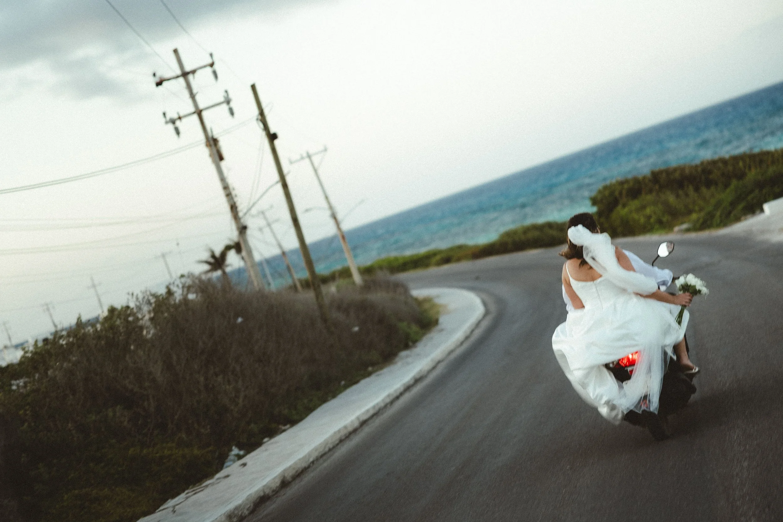 A bride in a white wedding dress riding a motorcycle on a highway near the coast, with the ocean and cloudy sky in the background.