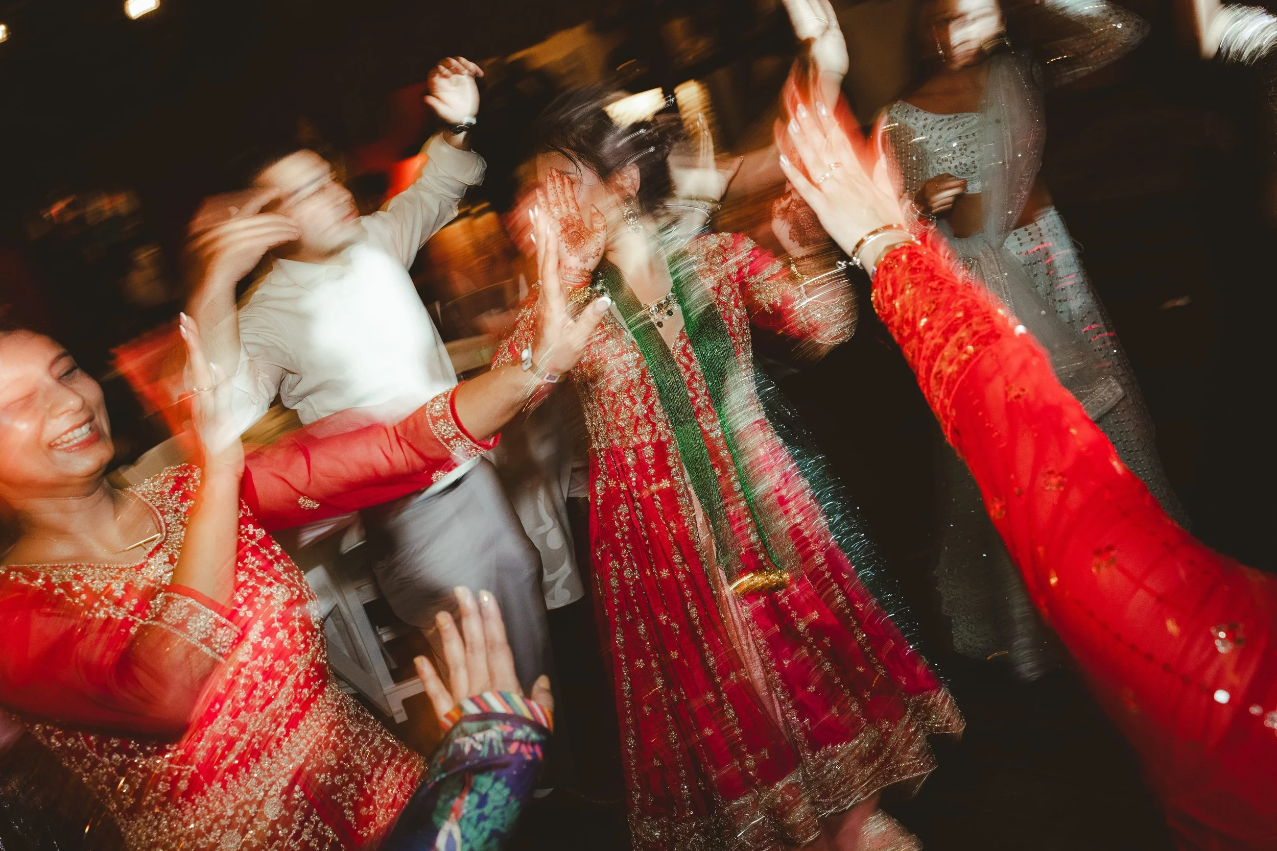 People dancing at a celebration, wearing festive Indian clothing, with some in sequined and embroidered outfits, captured in motion.