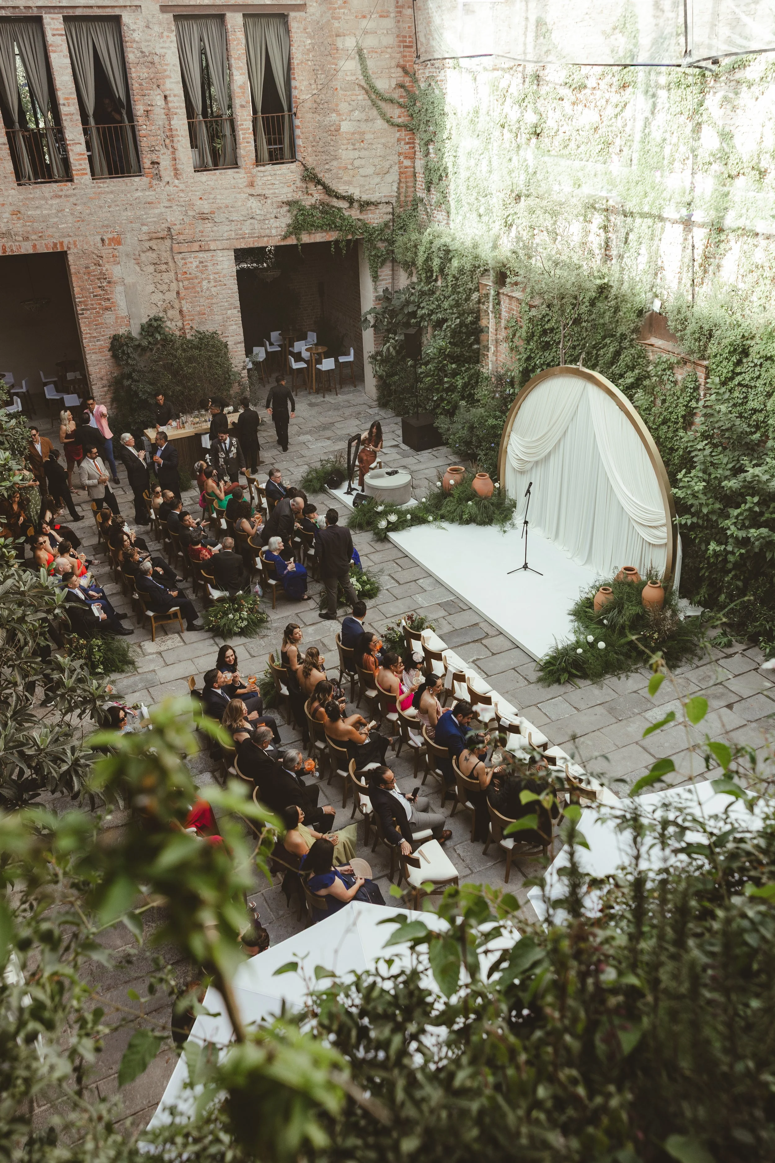 Indoor event taking place in a spacious, rustic industrial-style venue with brick walls and large windows. A crowd of people is seated and standing, watching a stage area decorated with greenery, large pots, and a white curtain backdrop. The stage ha
