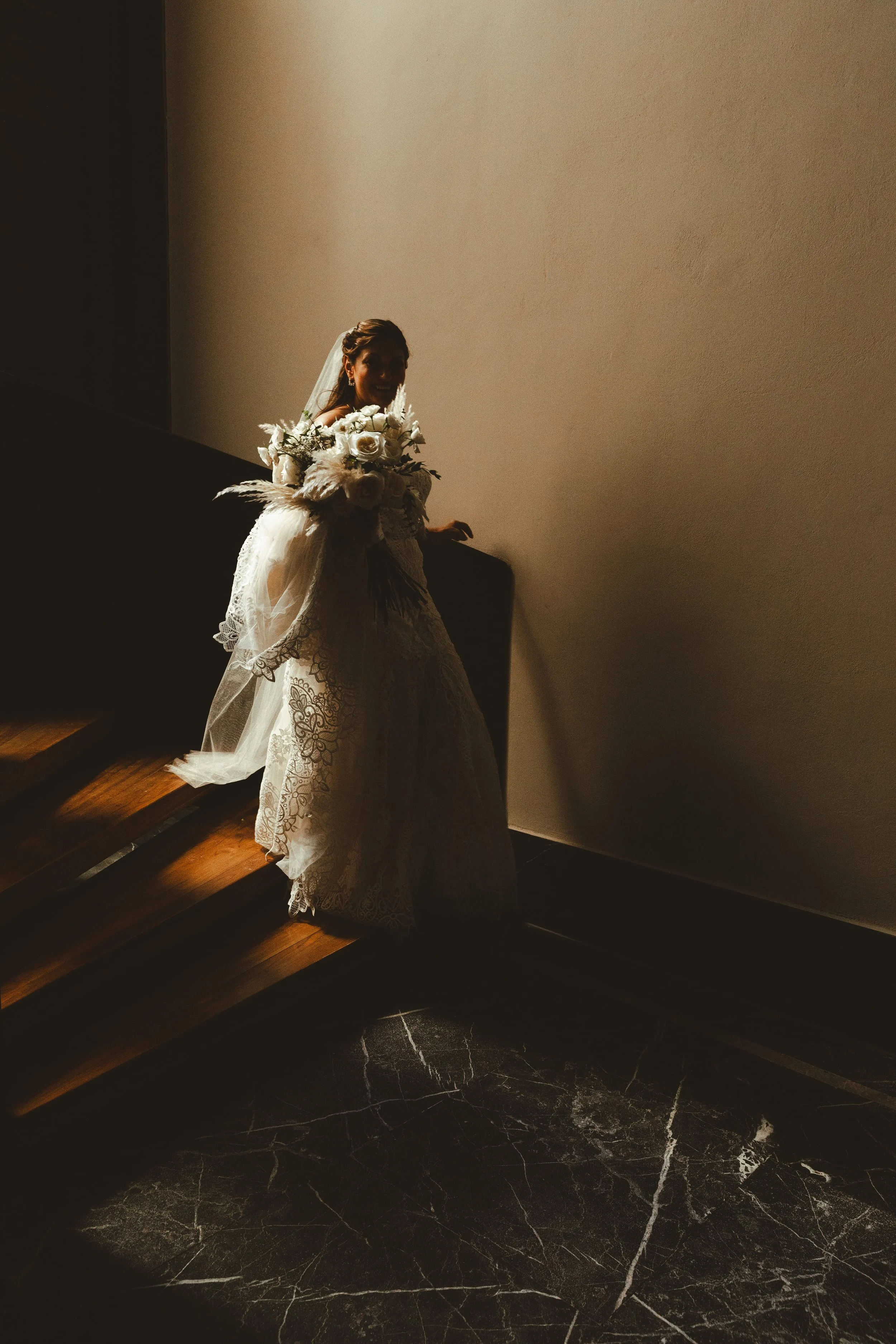 A bride holding a bouquet of white roses and greenery, standing on a staircase with a shadowed face and a veil, partially obscured by shadow.