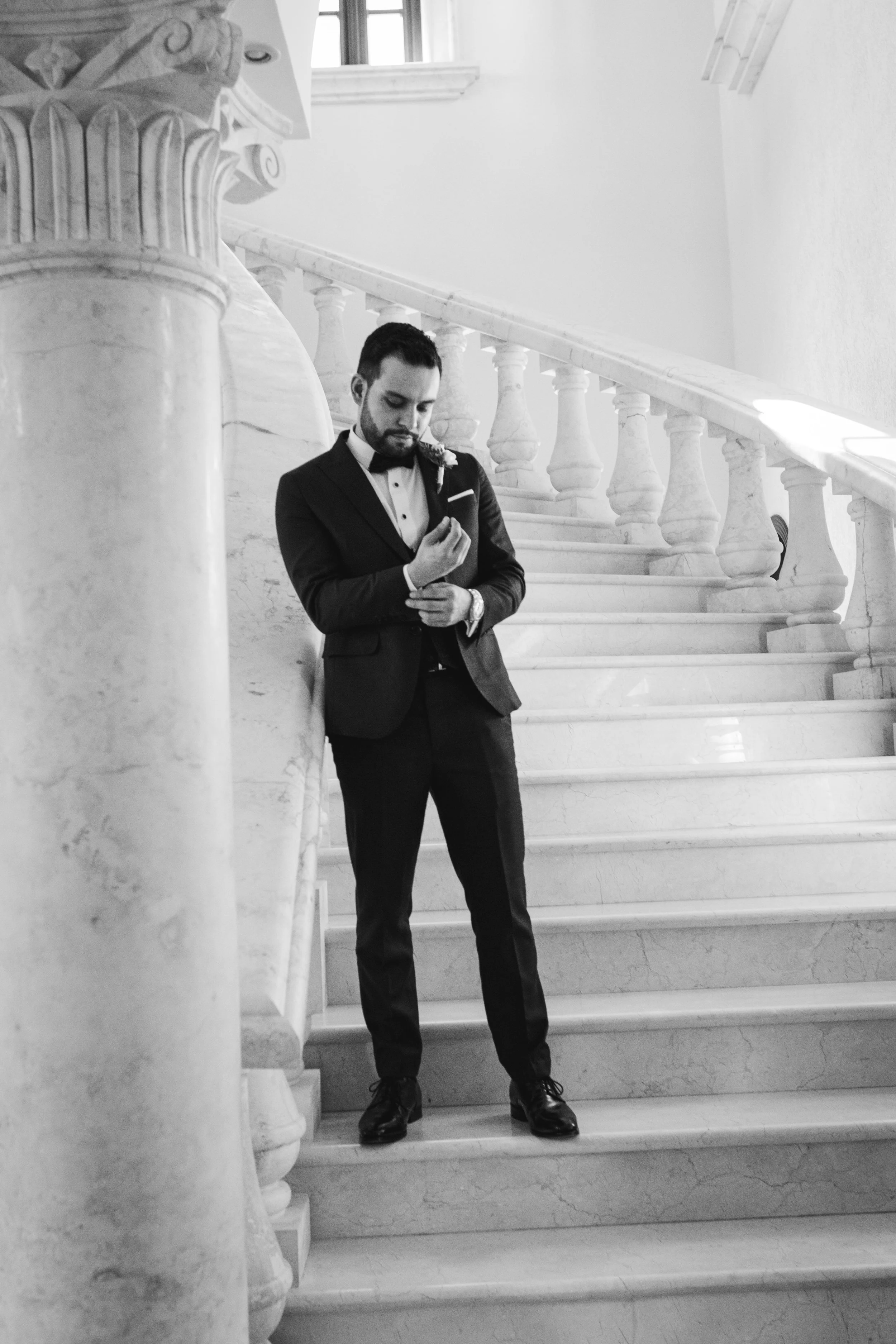 A groom in a tuxedo and bowtie standing on a marble staircase indoors, holding a flower, with a decorative marble railing and a window in the background.
