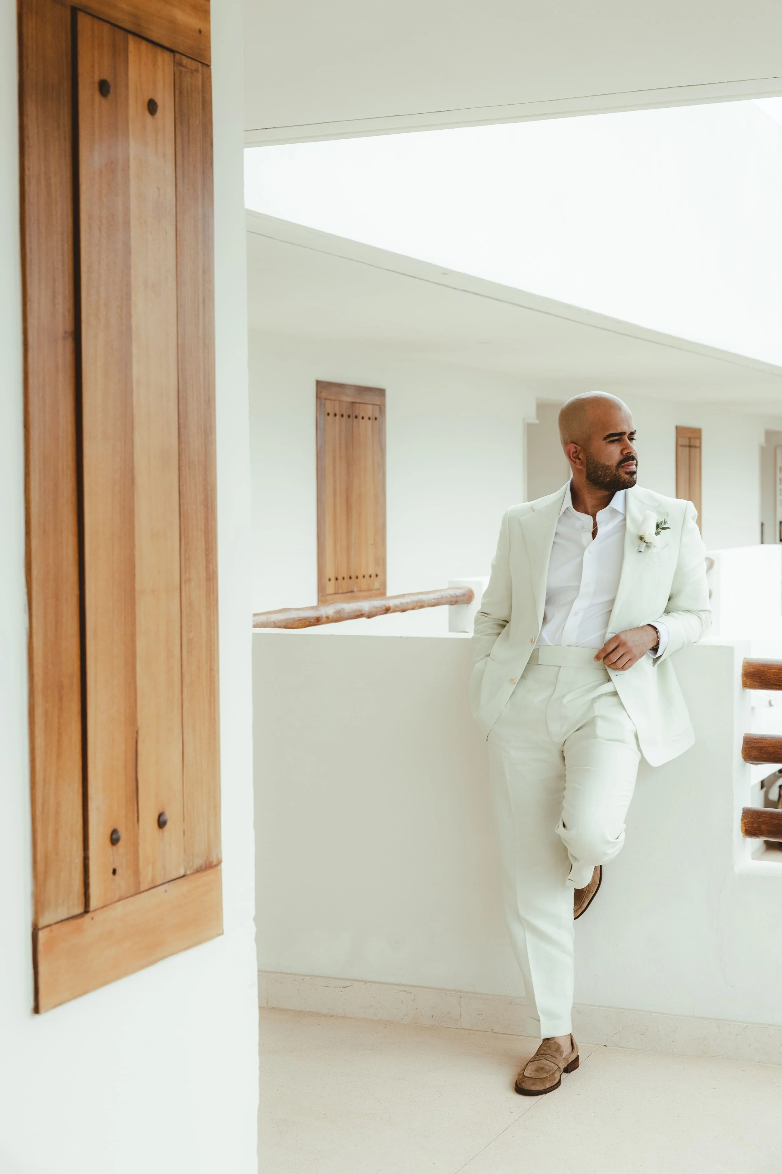 A man dressed in a white suit with a boutonnière, white shirt, and light brown shoes, leaning against a white railing in a modern, bright interior with wooden accents and stairs, looking to the side.