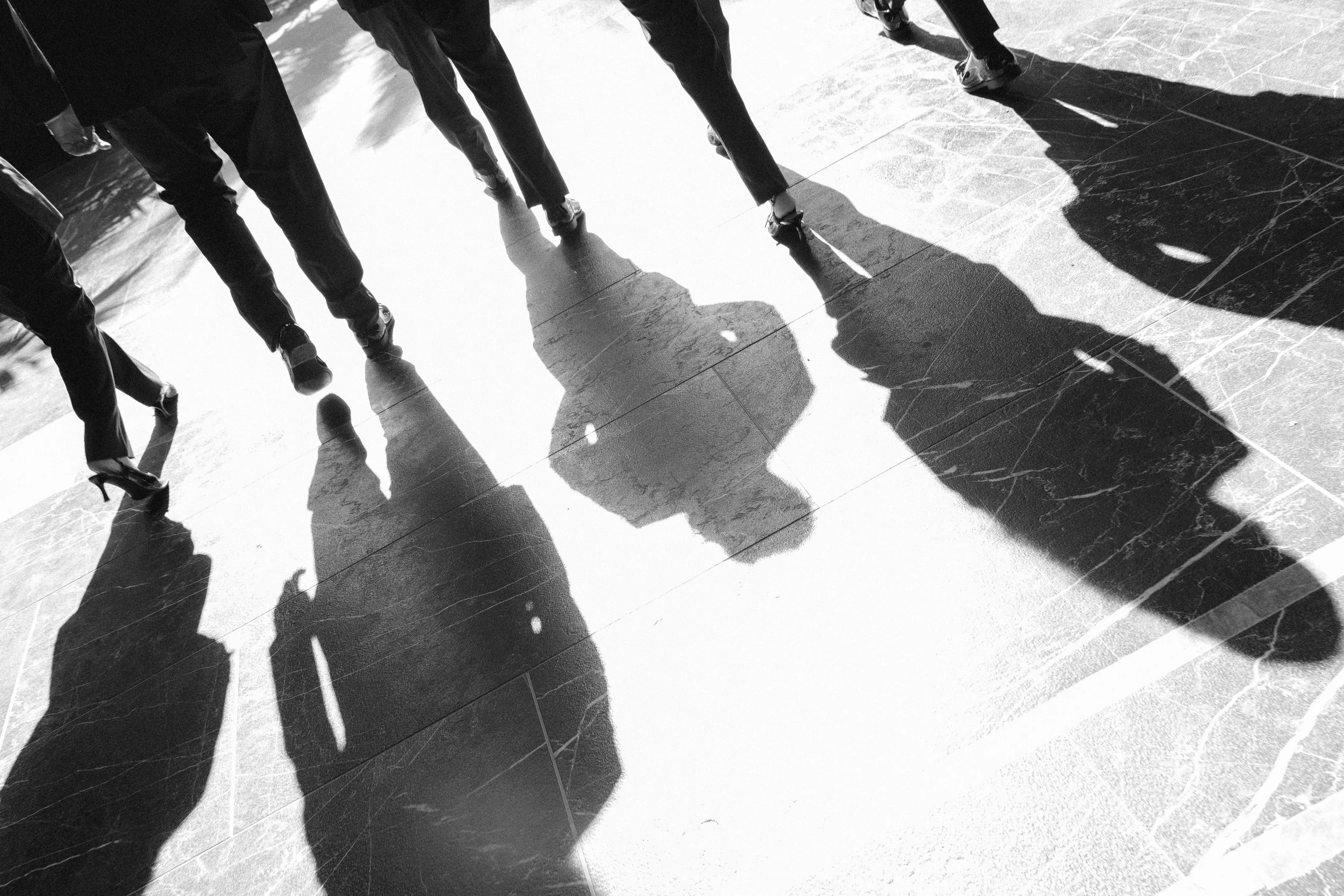 Shadows of people walking on a tiled floor, with sunlight casting shadows on the ground.