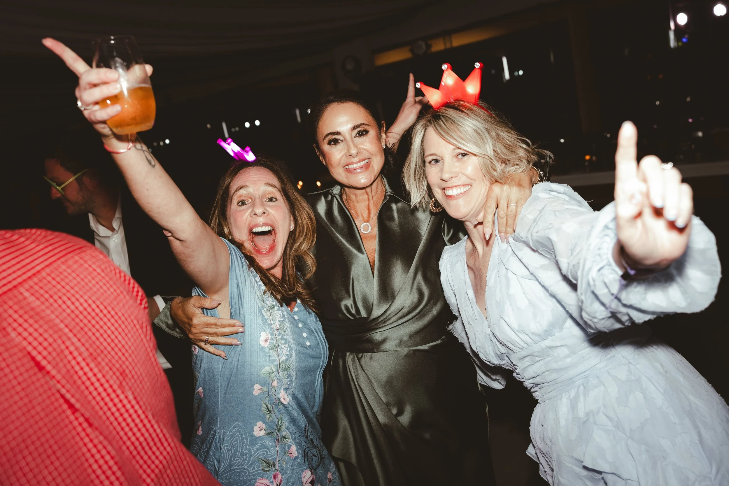 Group of women celebrating at a party, some wearing illuminated birthday crown accessories and holding drinks, smiling and posing for the camera.