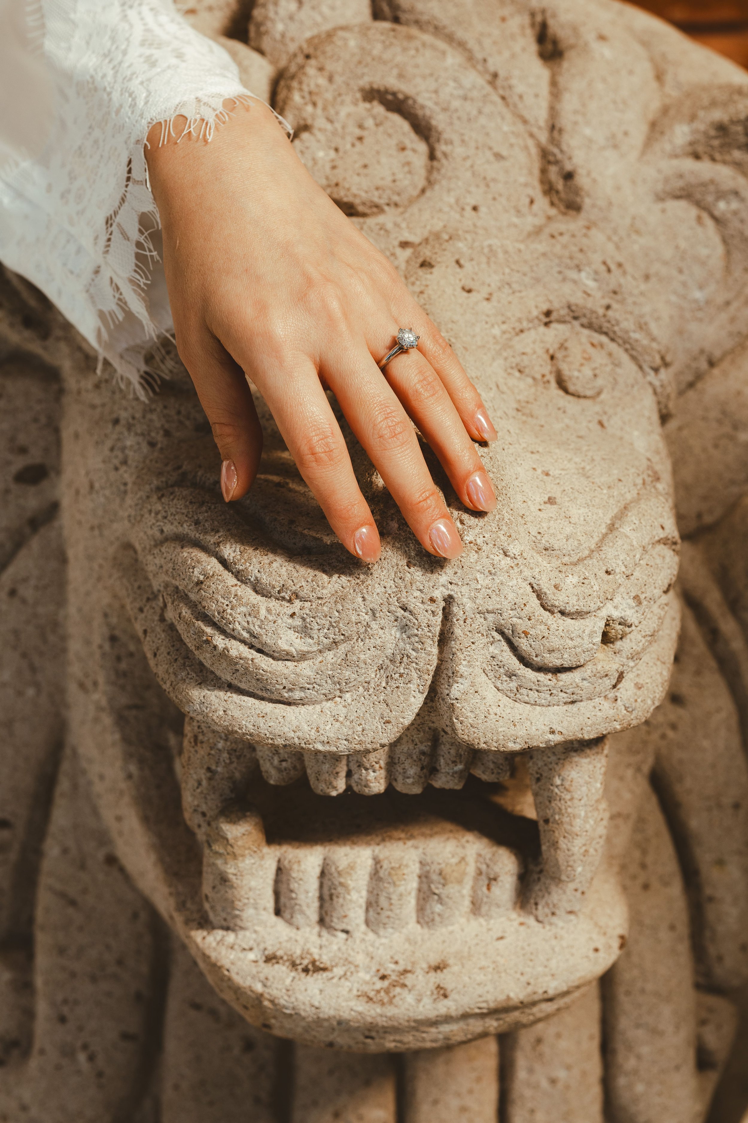 A hand with a diamond engagement ring rests on a carved stone sculpture of a face.