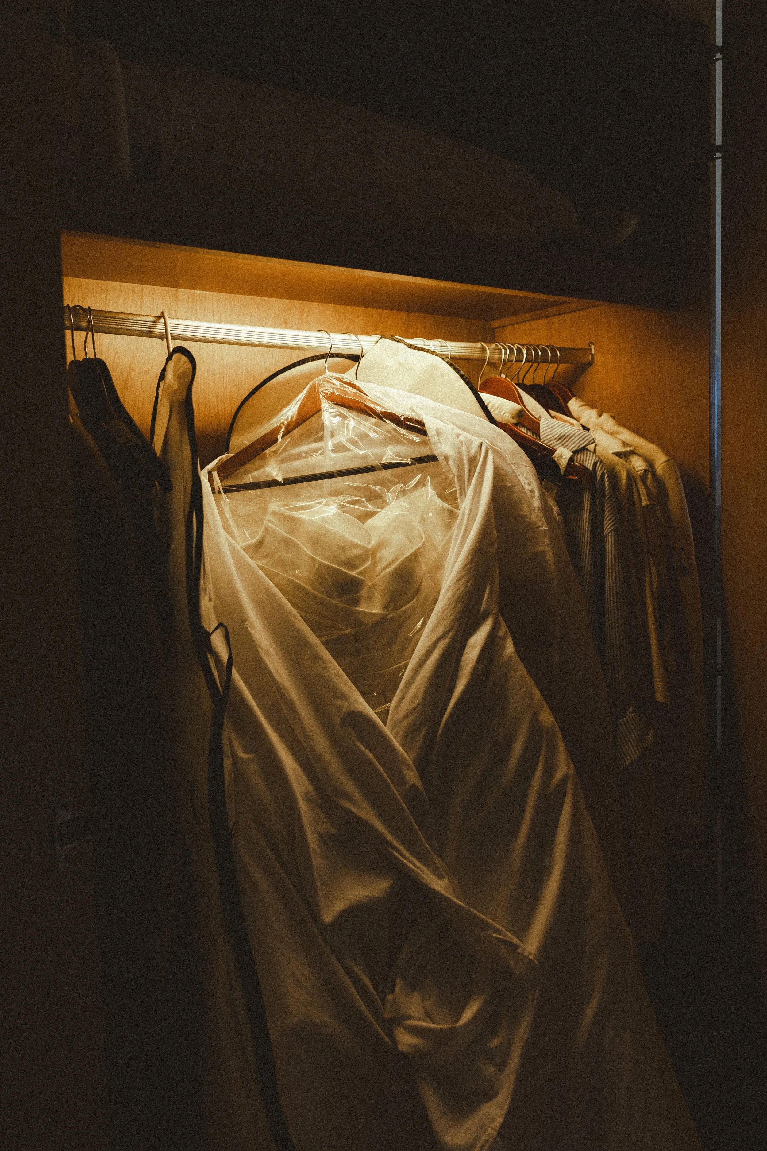 A wardrobe with various clothing items hanging on a metal rod inside a dimly lit closet.