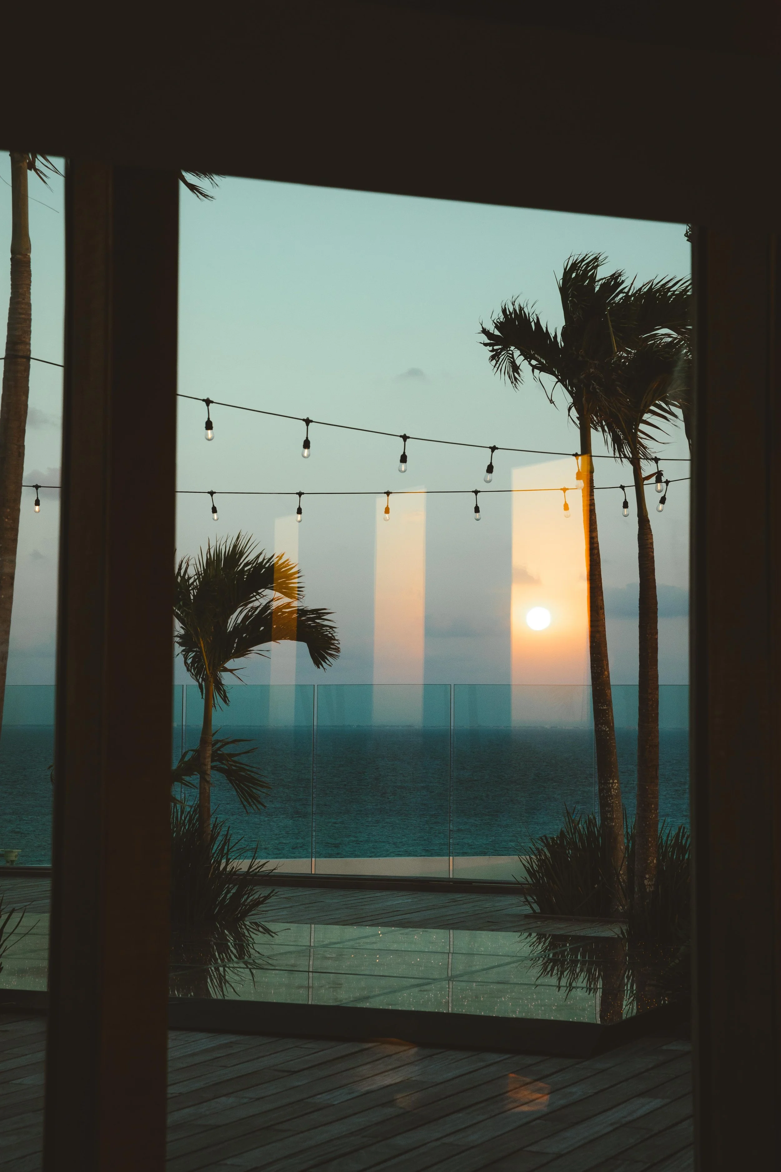 Sunset view through a glass barrier with palm trees and outdoor string lights, seen from a shaded indoor area.