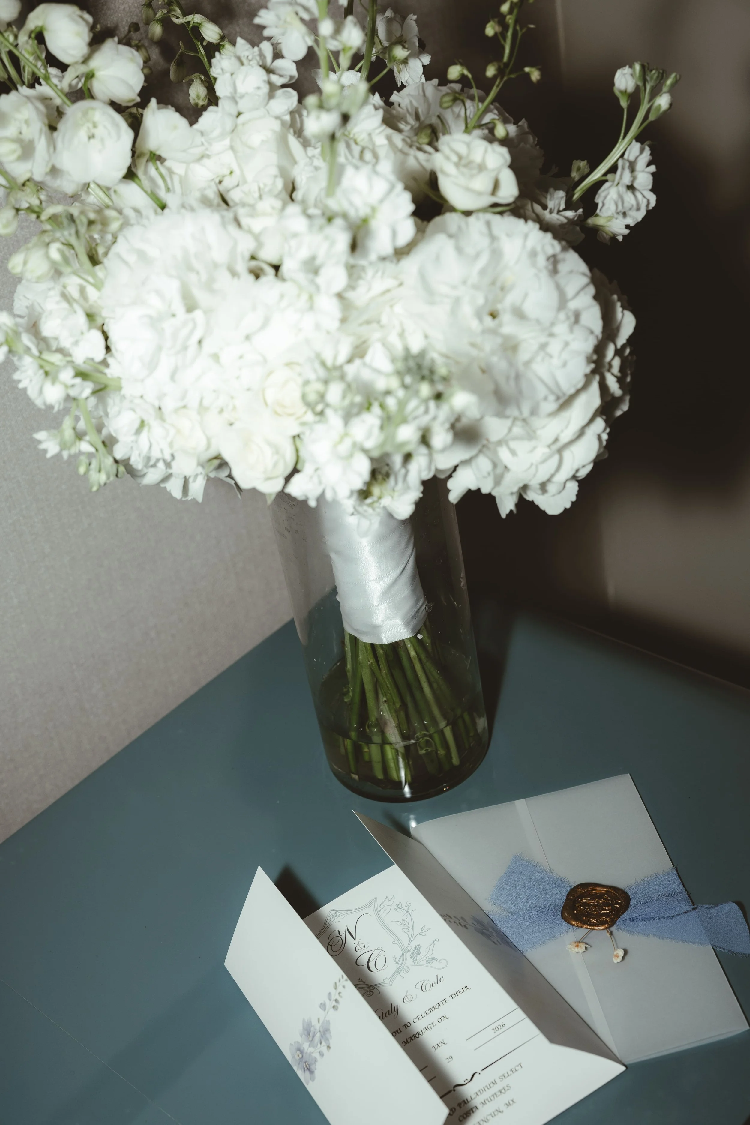 A bouquet of white flowers in a tall glass vase, with wedding invitation and envelope on a table.