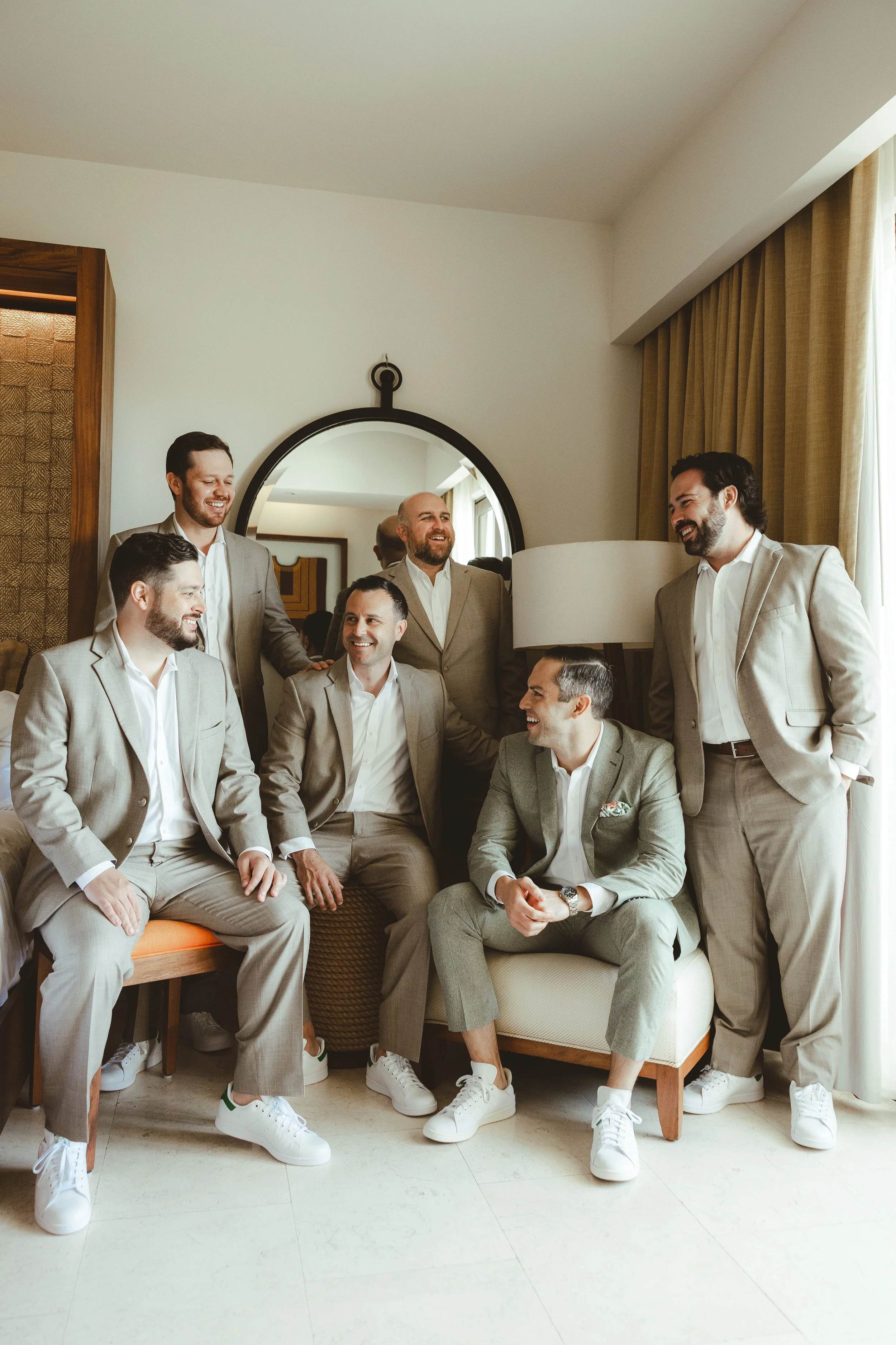 A group of eight men in light-colored suits and white sneakers, posed in a living room with neutral walls, a round mirror on the wall, and beige curtains. They are smiling and engaging in conversation.