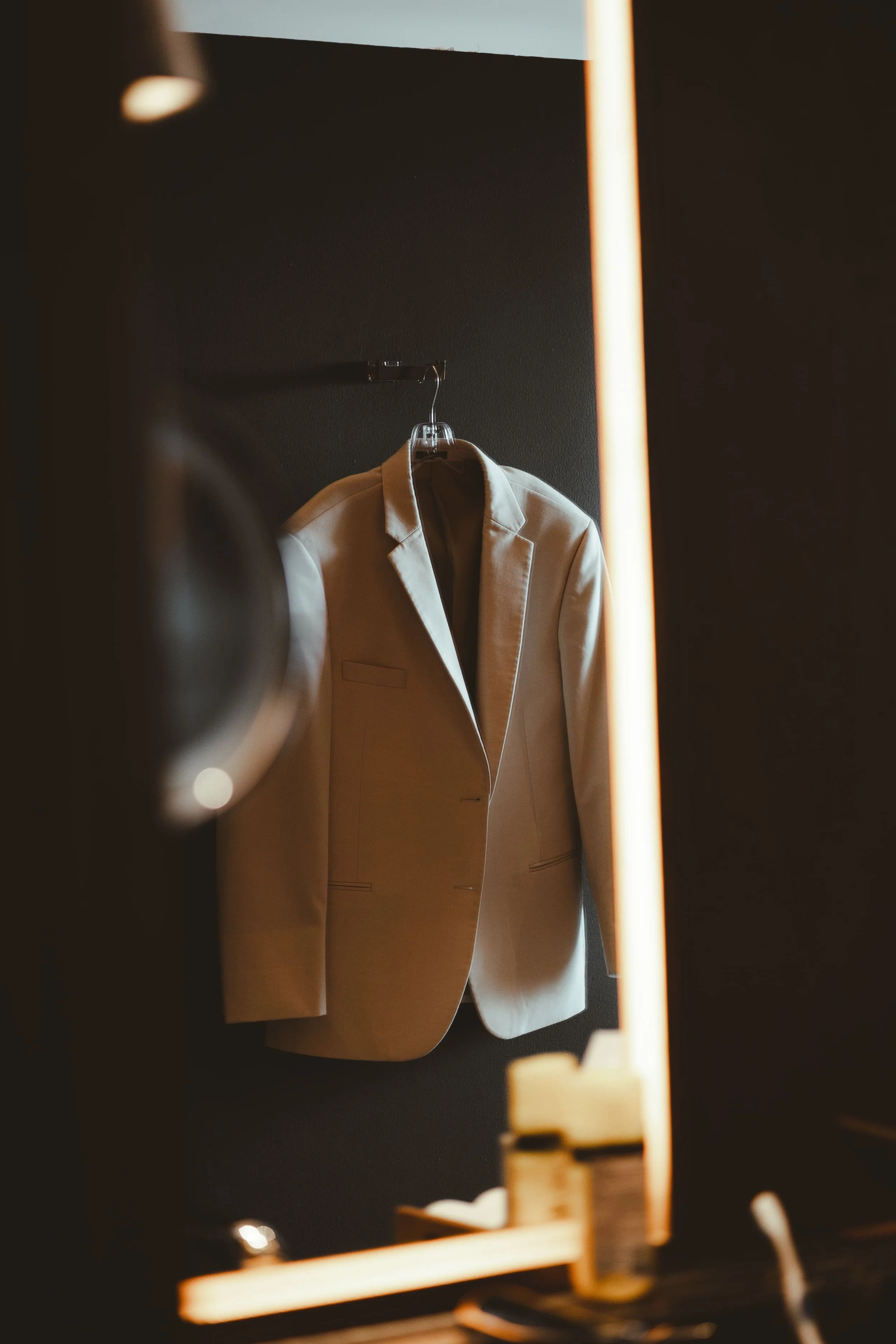 A beige blazer hangs on a black hanger against a dark background, with warm lighting creating shadows.