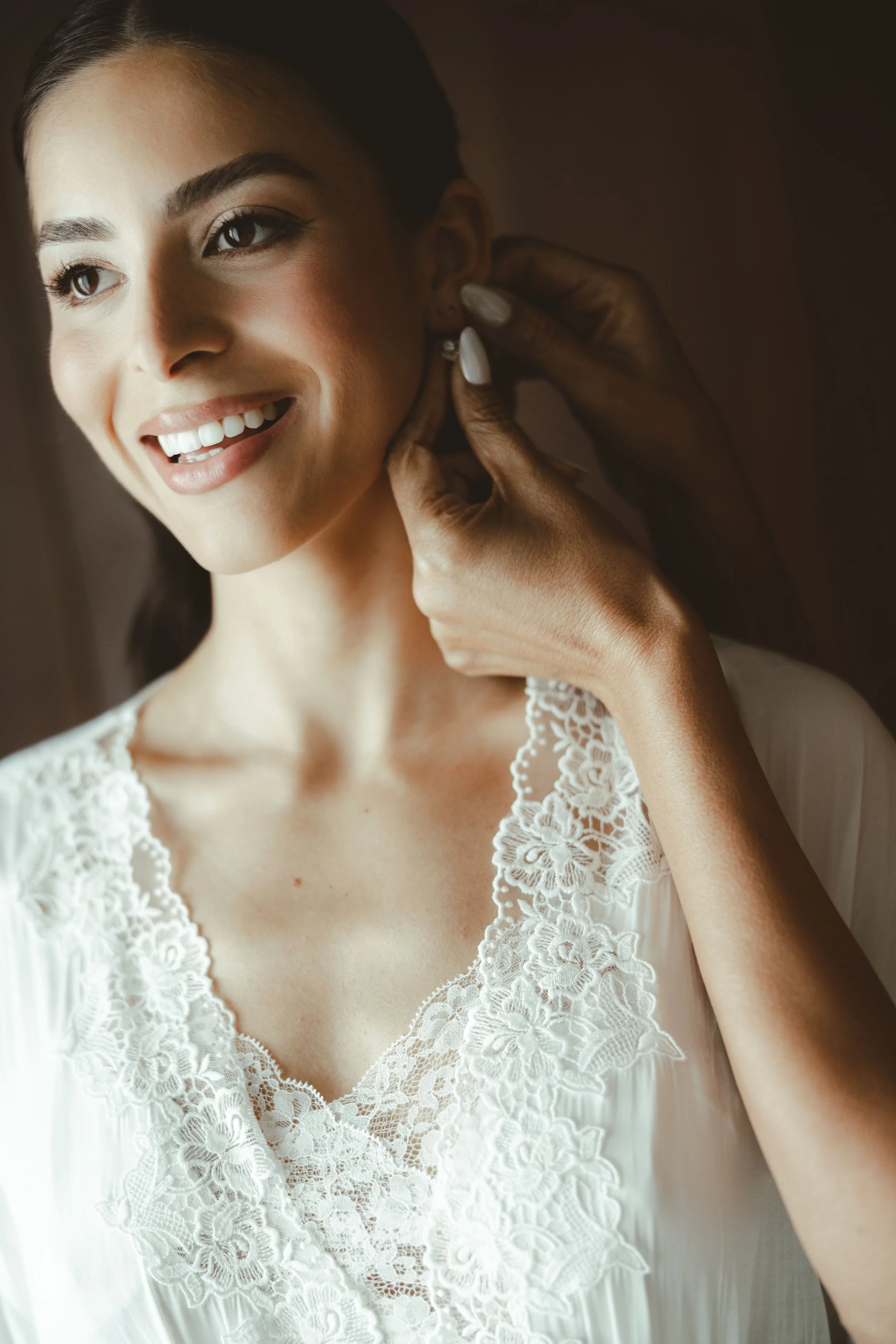 A woman smiling as she puts on an earring, wearing a white lace dress.