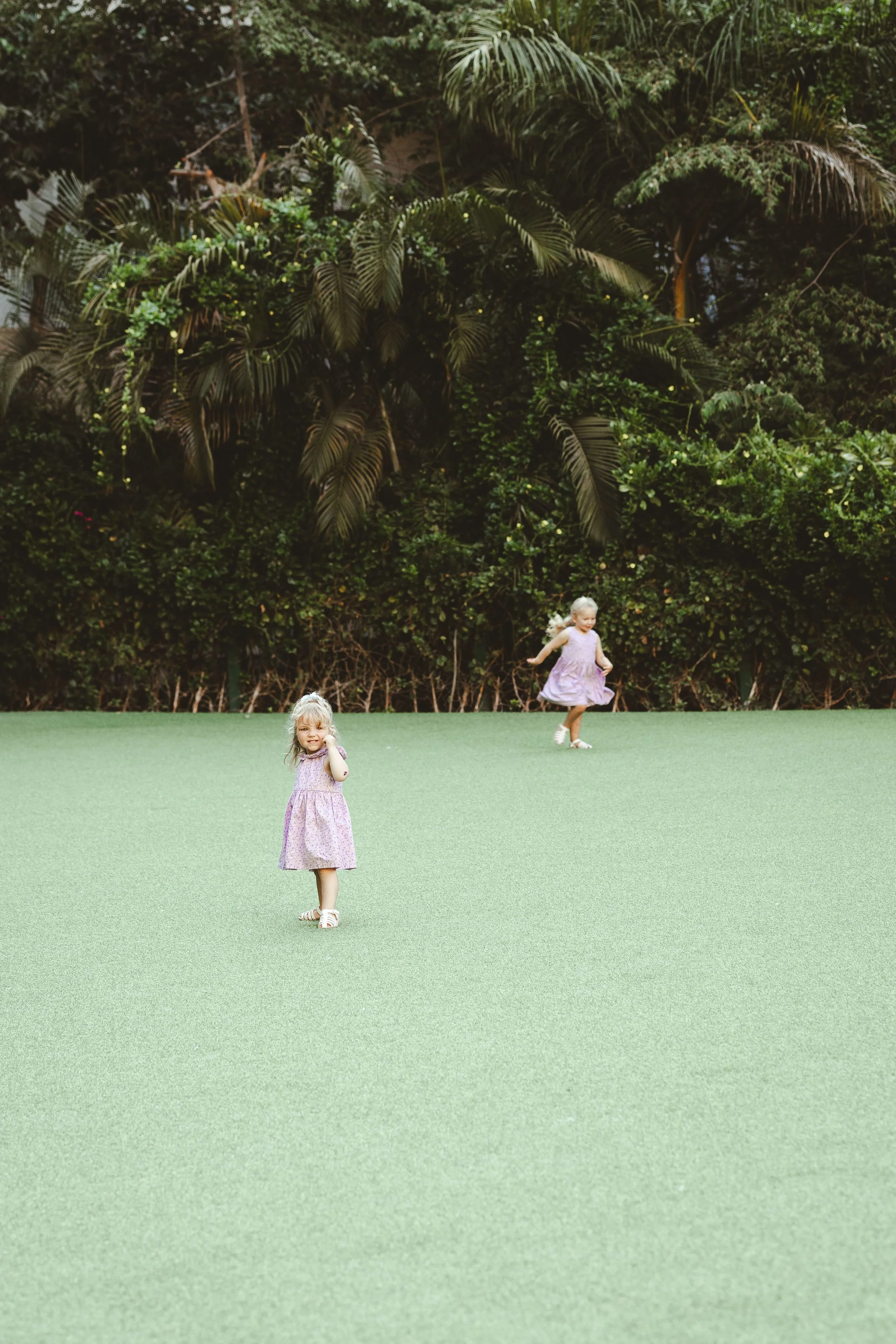 Two young girls in pink dresses playing on a green artificial lawn with a dense background of jungle-like trees and shrubs.