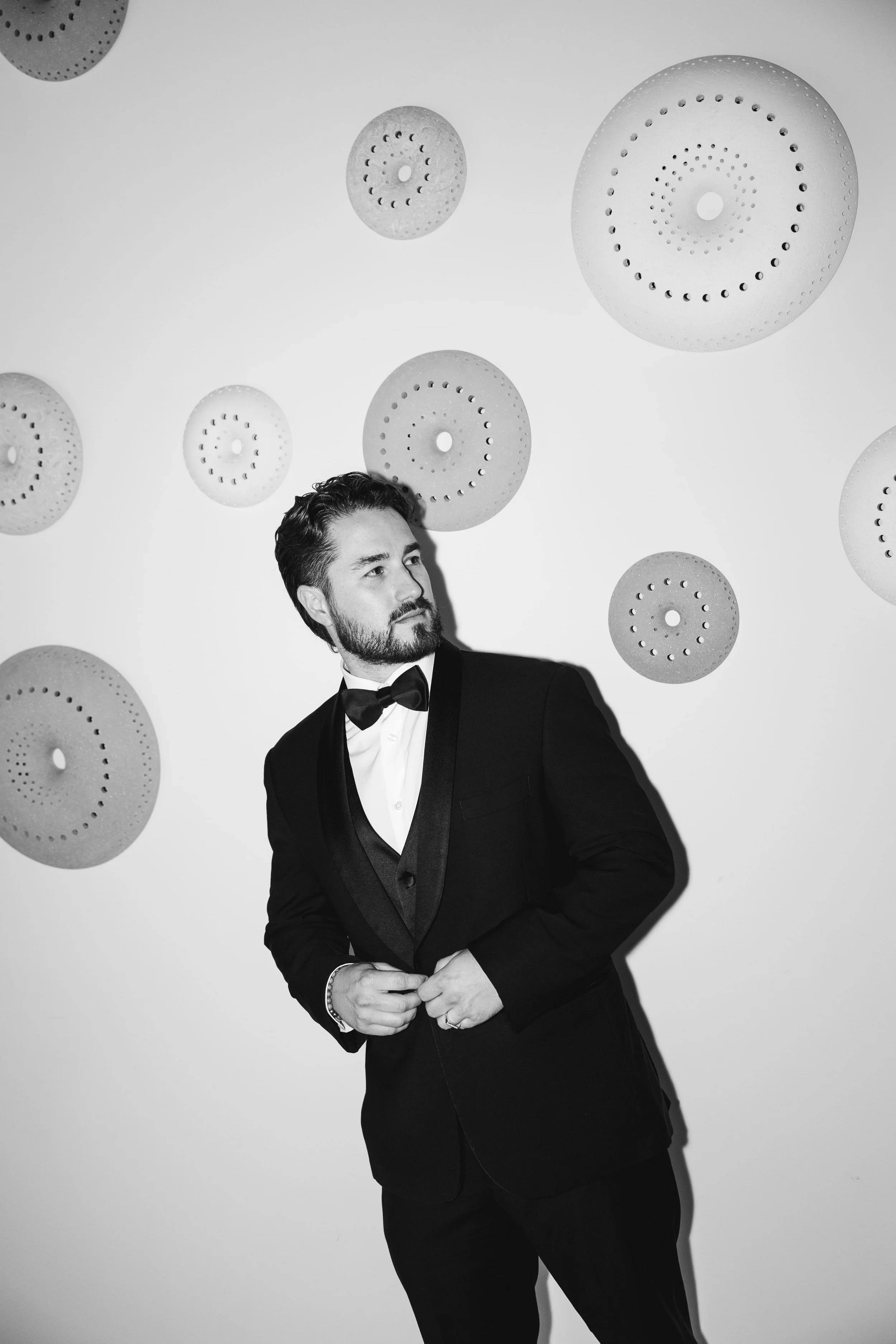 A man dressed in a tuxedo with a bow tie standing against a white wall decorated with circular lighting fixtures.