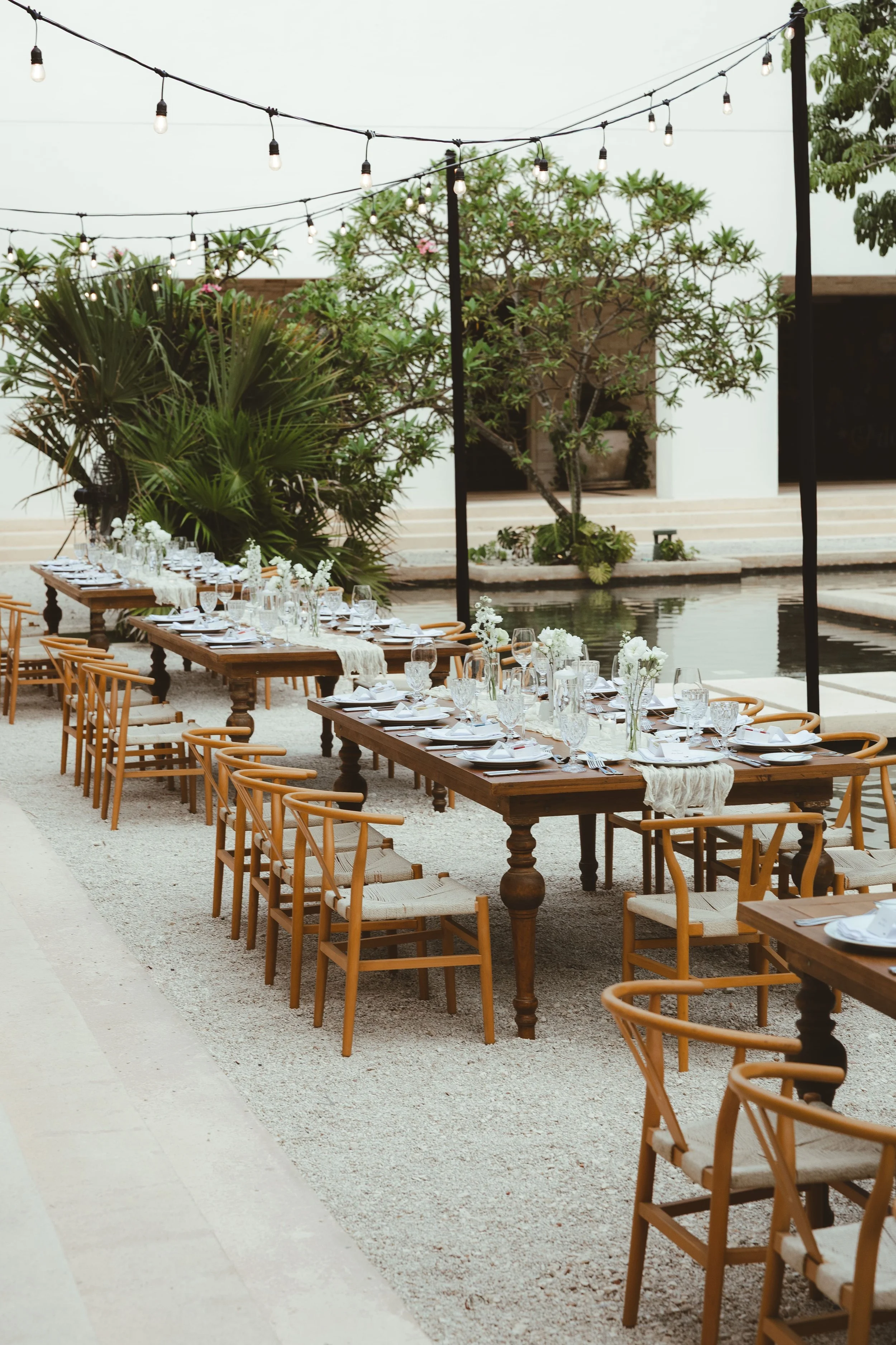 Outdoor wedding dining setup with long wooden tables, chairs, and white floral centerpieces. String lights hang overhead, with greenery and trees in the background near water. Secrets Akumal.