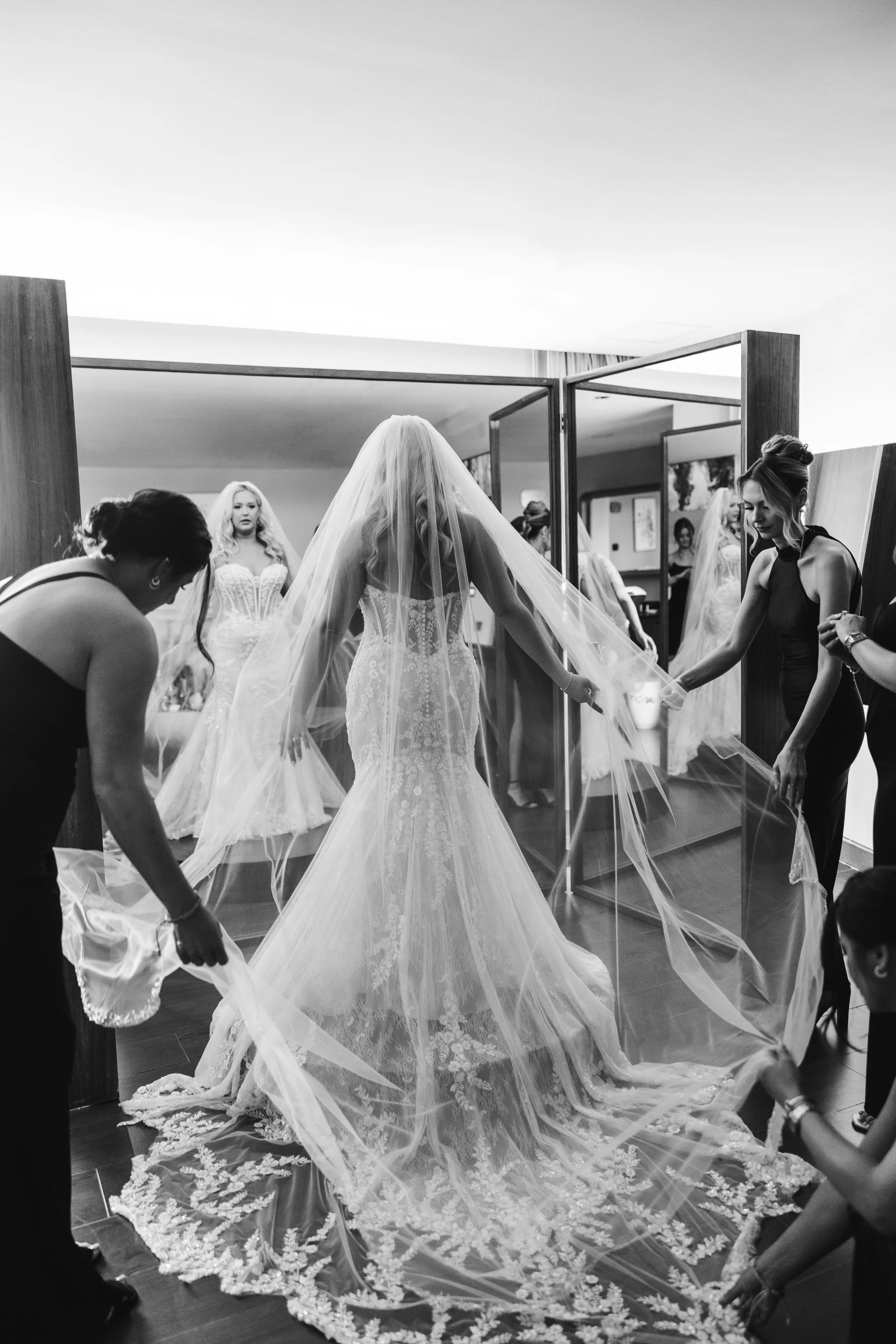 Women assisting a bride with her wedding dress and veil in a bridal preparation room.