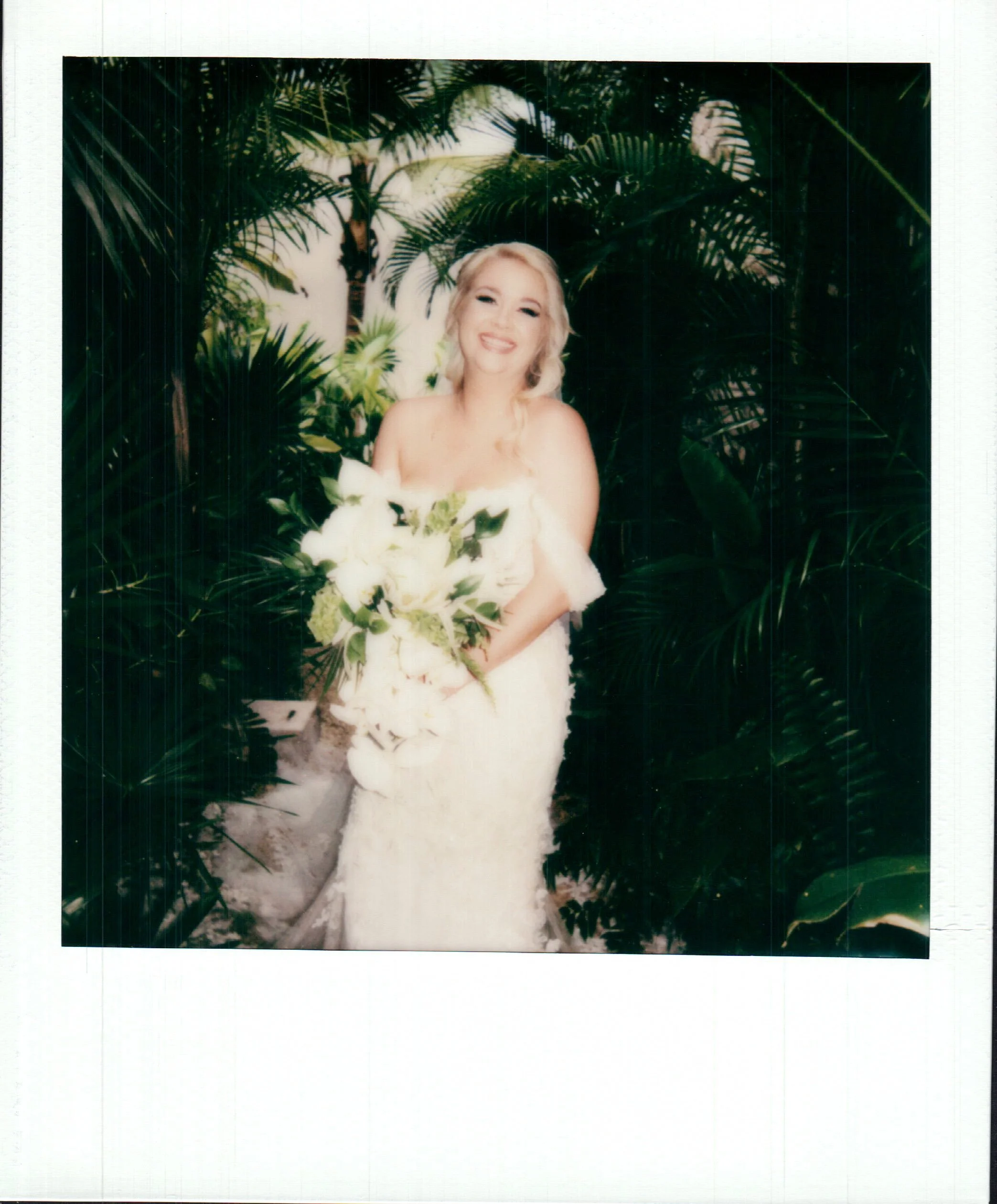 A woman with blonde hair smiling and wearing a white dress with greenery, holding a bouquet of white flowers, standing in front of lush green tropical plants with a white background.