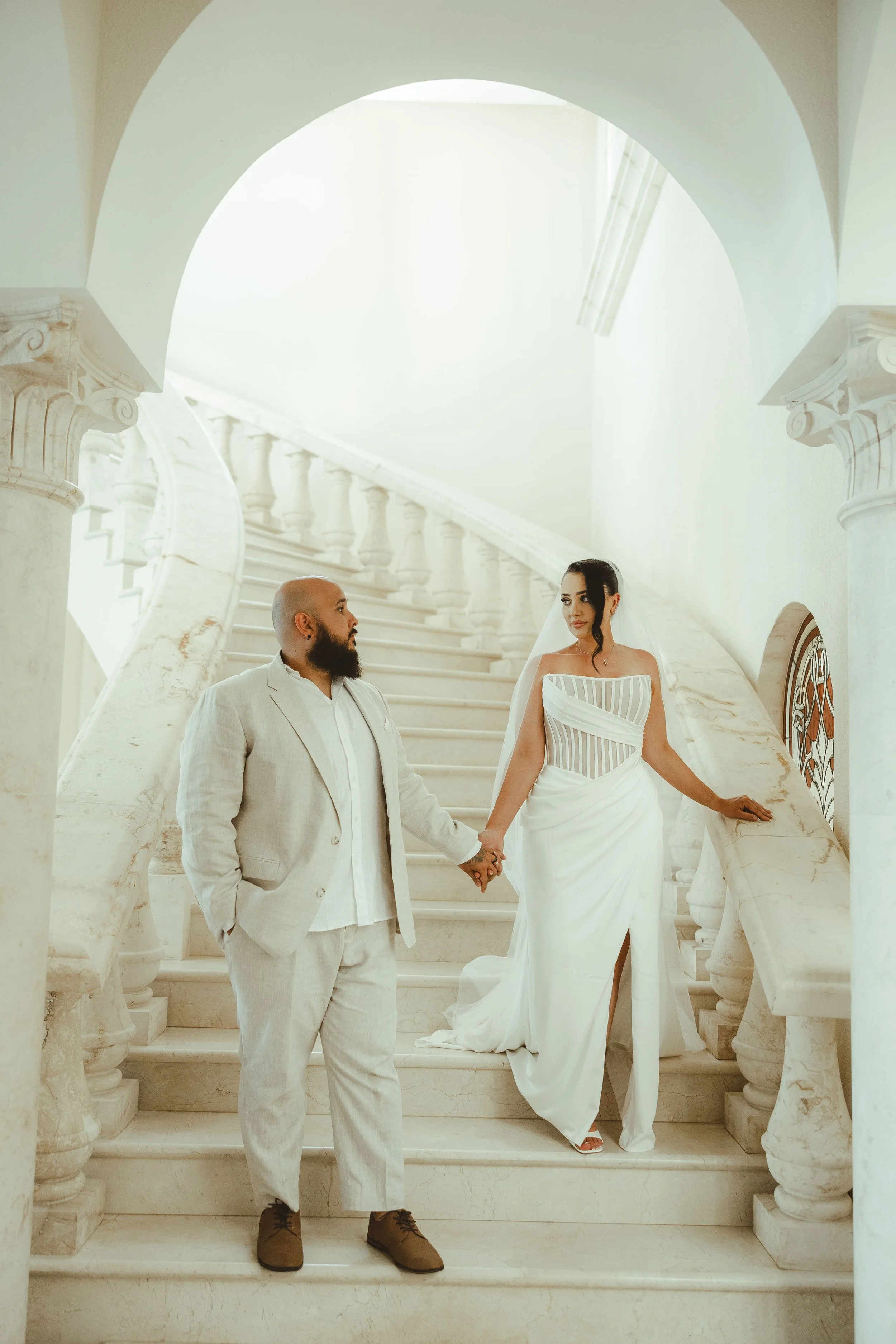 A bride and groom standing on a grand marble staircase, holding hands, in an elegant, well-lit interior with classical architecture and marble columns.