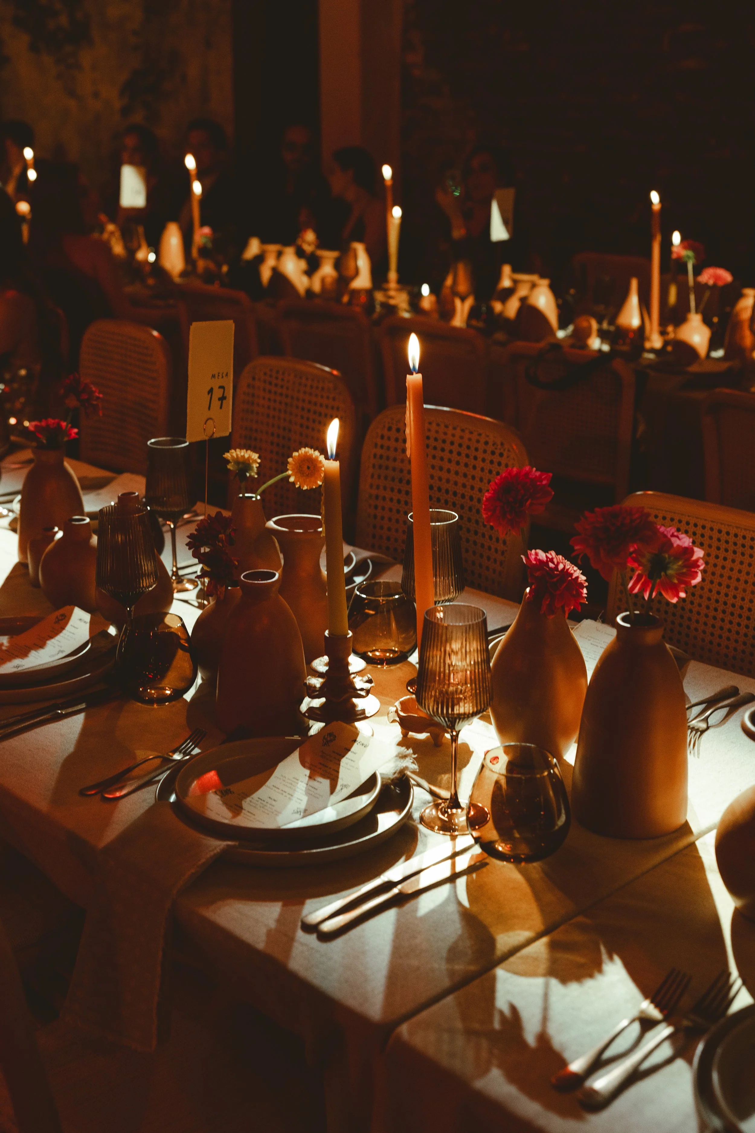 A dimly lit banquet table set with brown ceramic vases holding pink and red flowers, tall candles in holders providing warm light, wine glasses, dinner plates with menus, cutlery, and table numbers, with people seated in the background in a rustic se