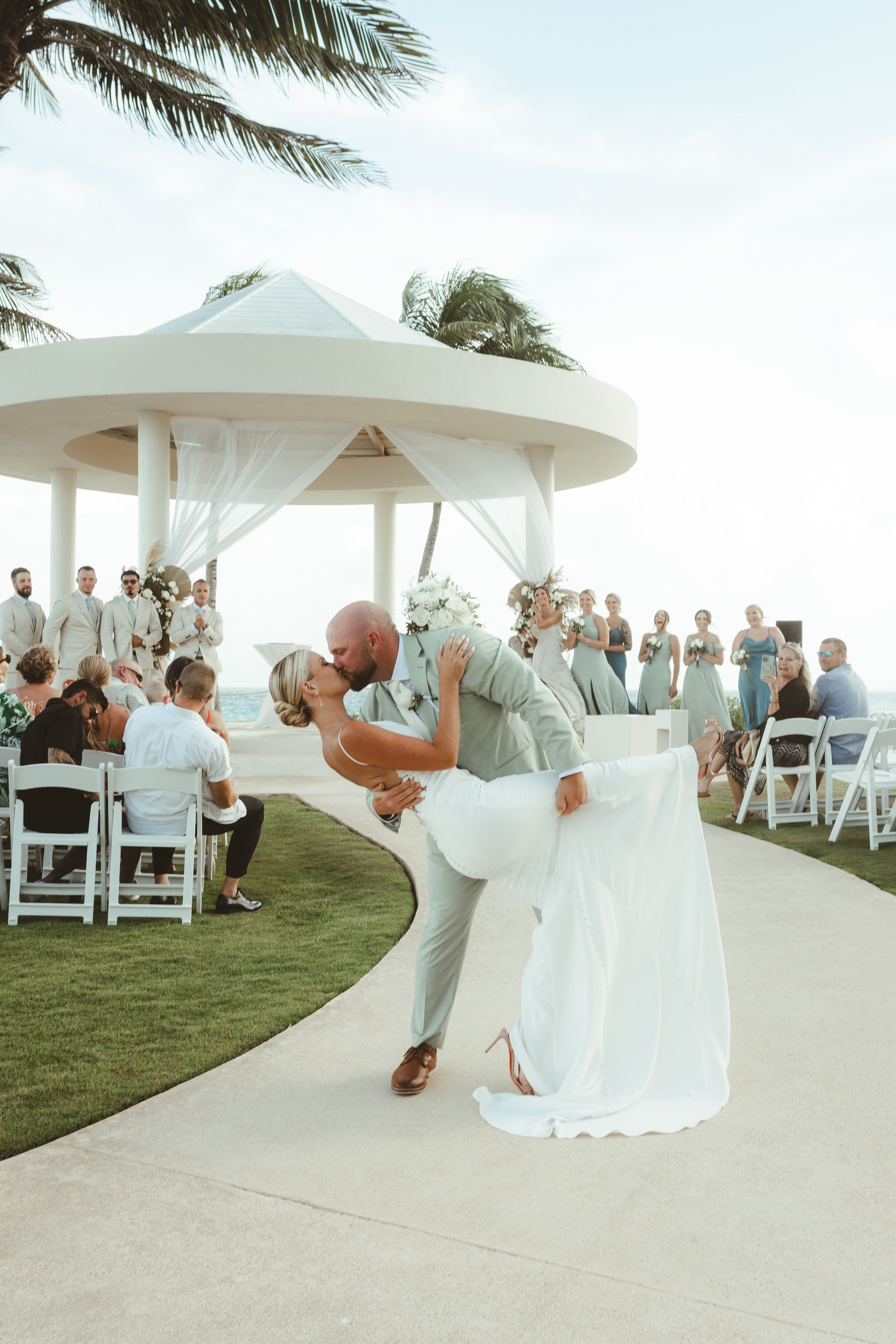 A wedding ceremony on the beach with the bride and groom sharing a kiss, surrounded by guests and wedding party, under a white canopy with ocean view in Hyatt Ziva Cancun.