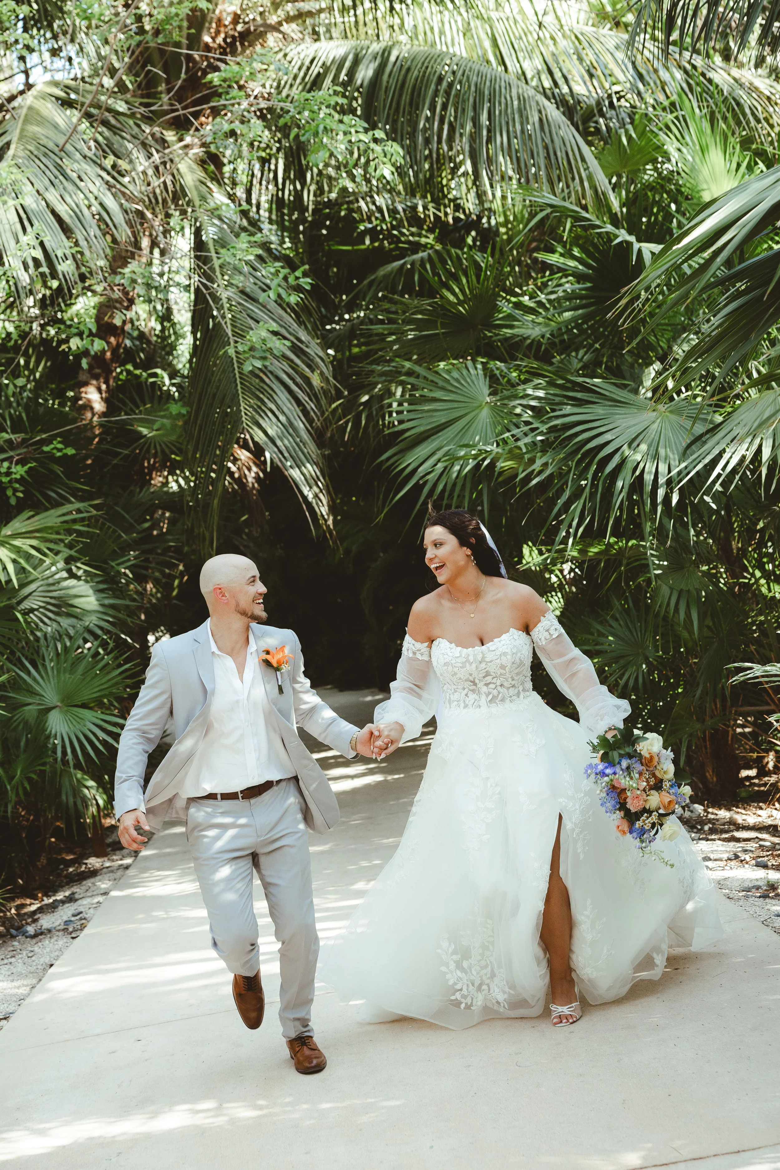 A bride and groom holding hands and walking outdoors on a sunny day, surrounded by lush green tropical plants, smiling at each other.