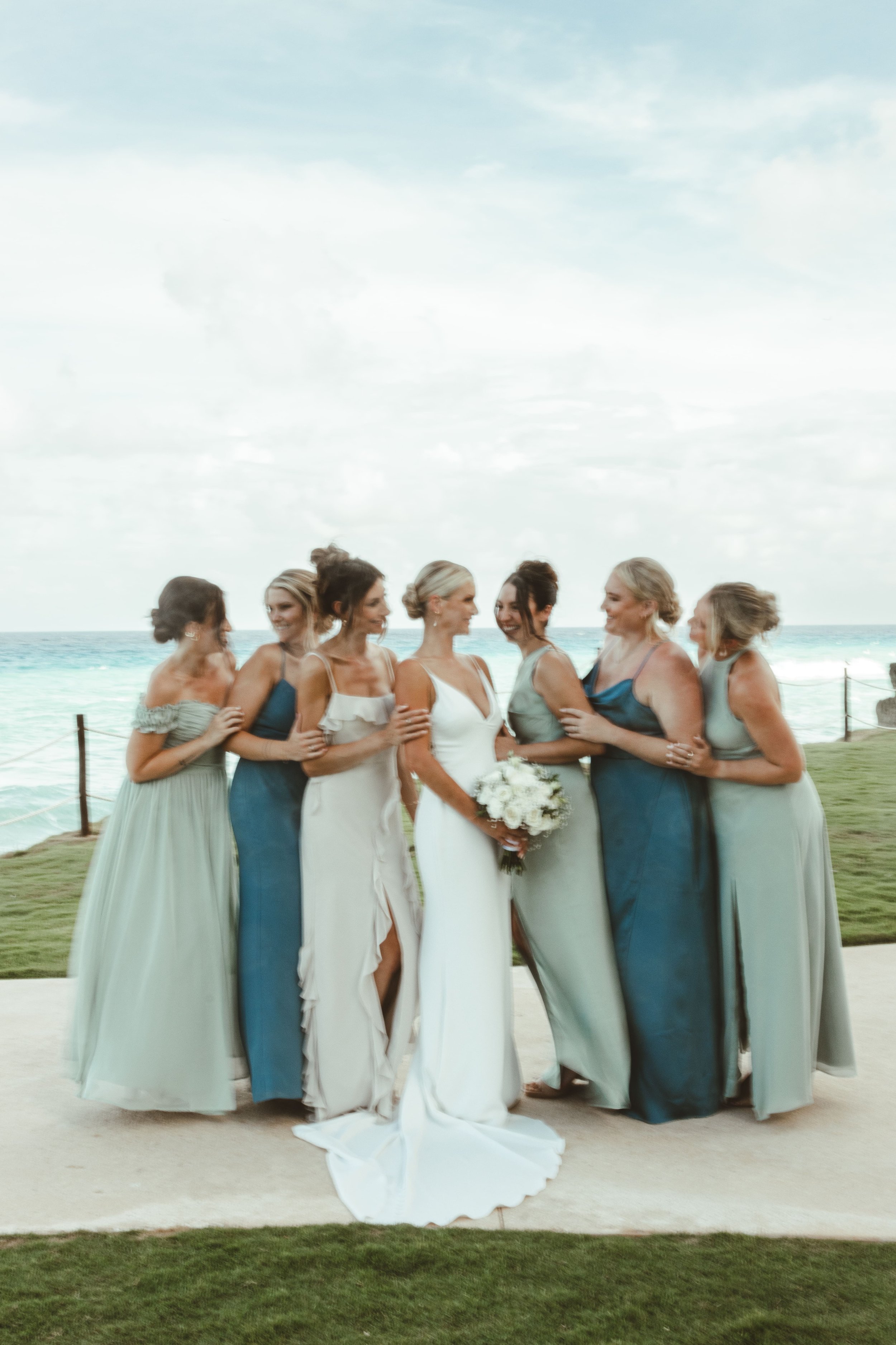 A group of women in formal dresses standing together on a beach, with the ocean and sky in the background, during a wedding or special event. Bridesmaids dresses are in blue and green color palette.