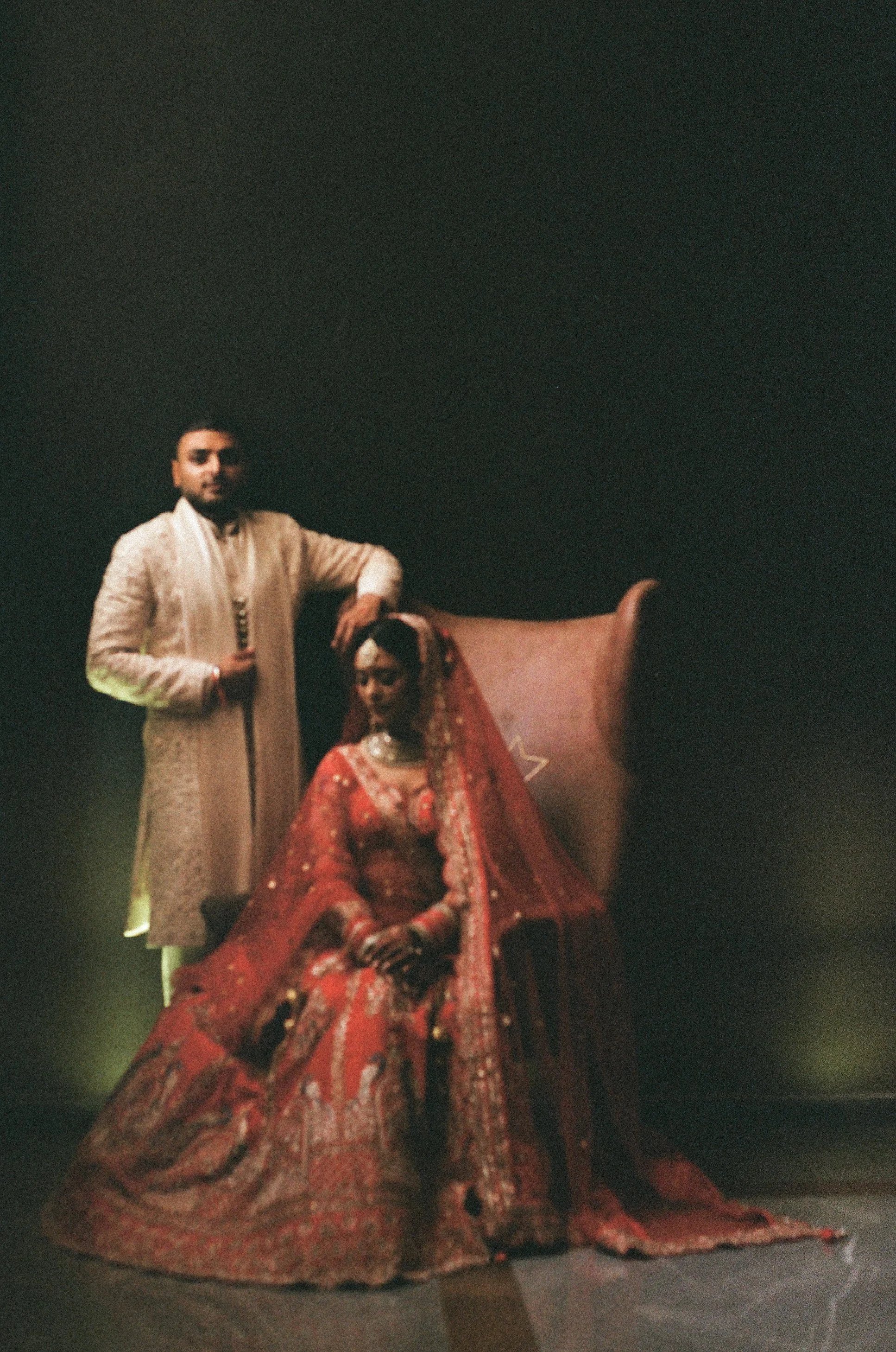 A man and a woman dressed in traditional wedding attire, standing in front of a dark background. The man is in a cream-colored sherwani with intricate embroidery, holding a gun, and has his arm resting on a chair. The woman is seated, wearing a red a