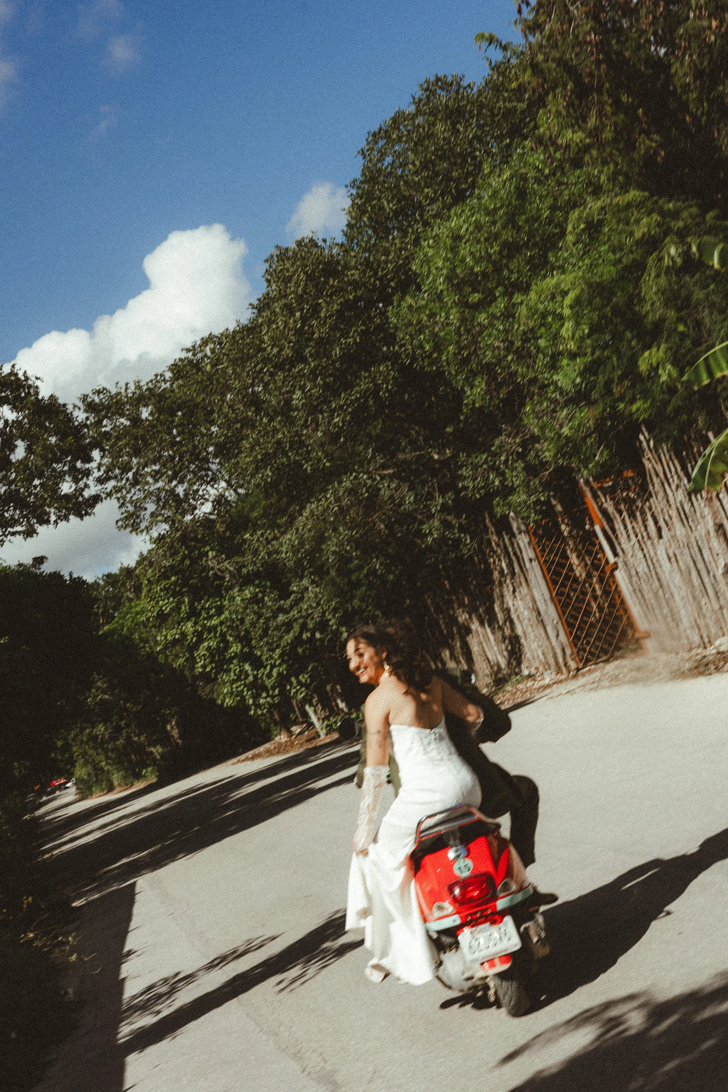A woman in a white dress riding a scooter on a road, with a smiling expression and trees in the background.