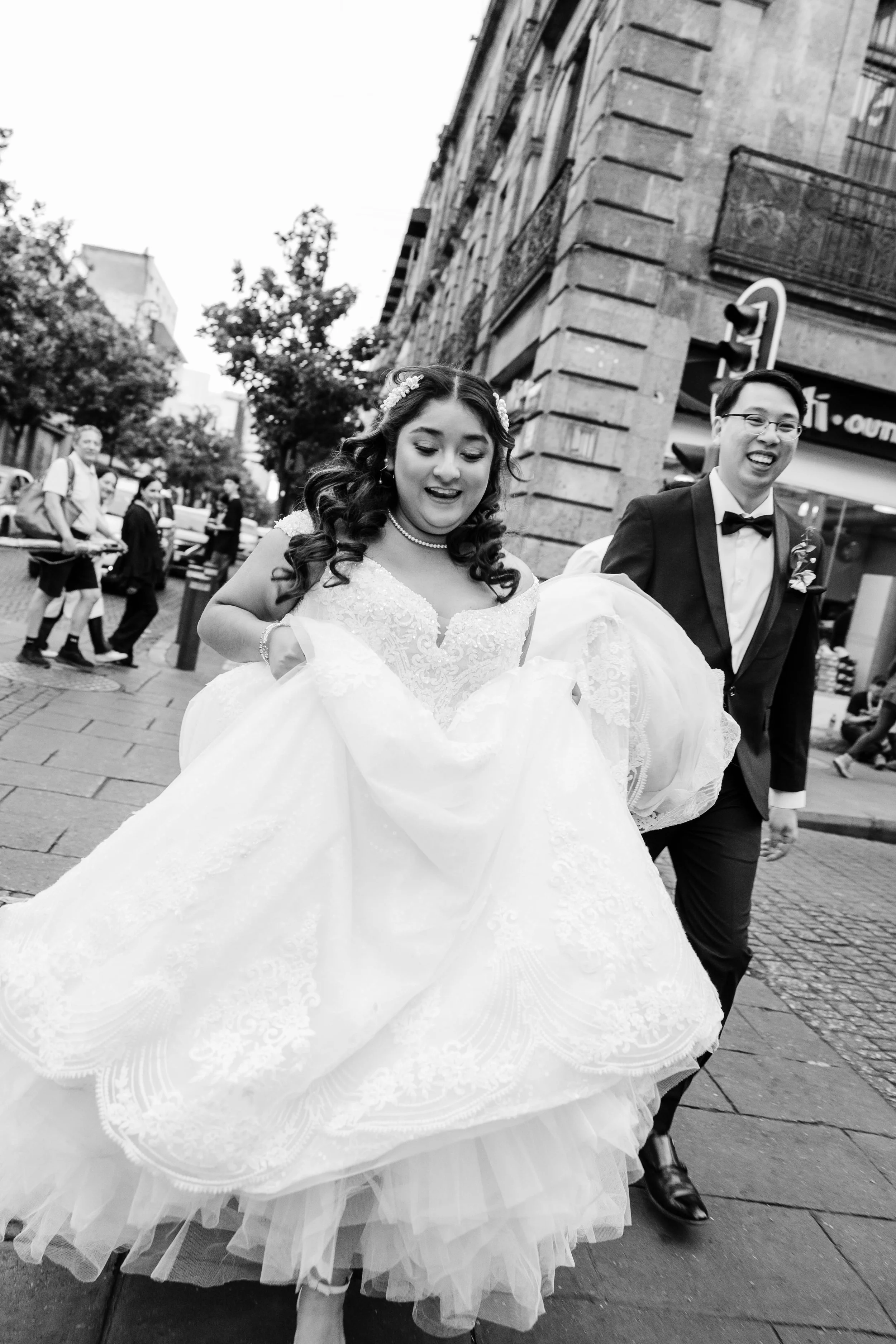 Black and white photo of a joyful bride and groom walking on a city sidewalk, with a group of people in the background. The bride is wearing a wedding gown with lace details and holding the skirt up slightly. The groom is dressed in a tuxedo with a b