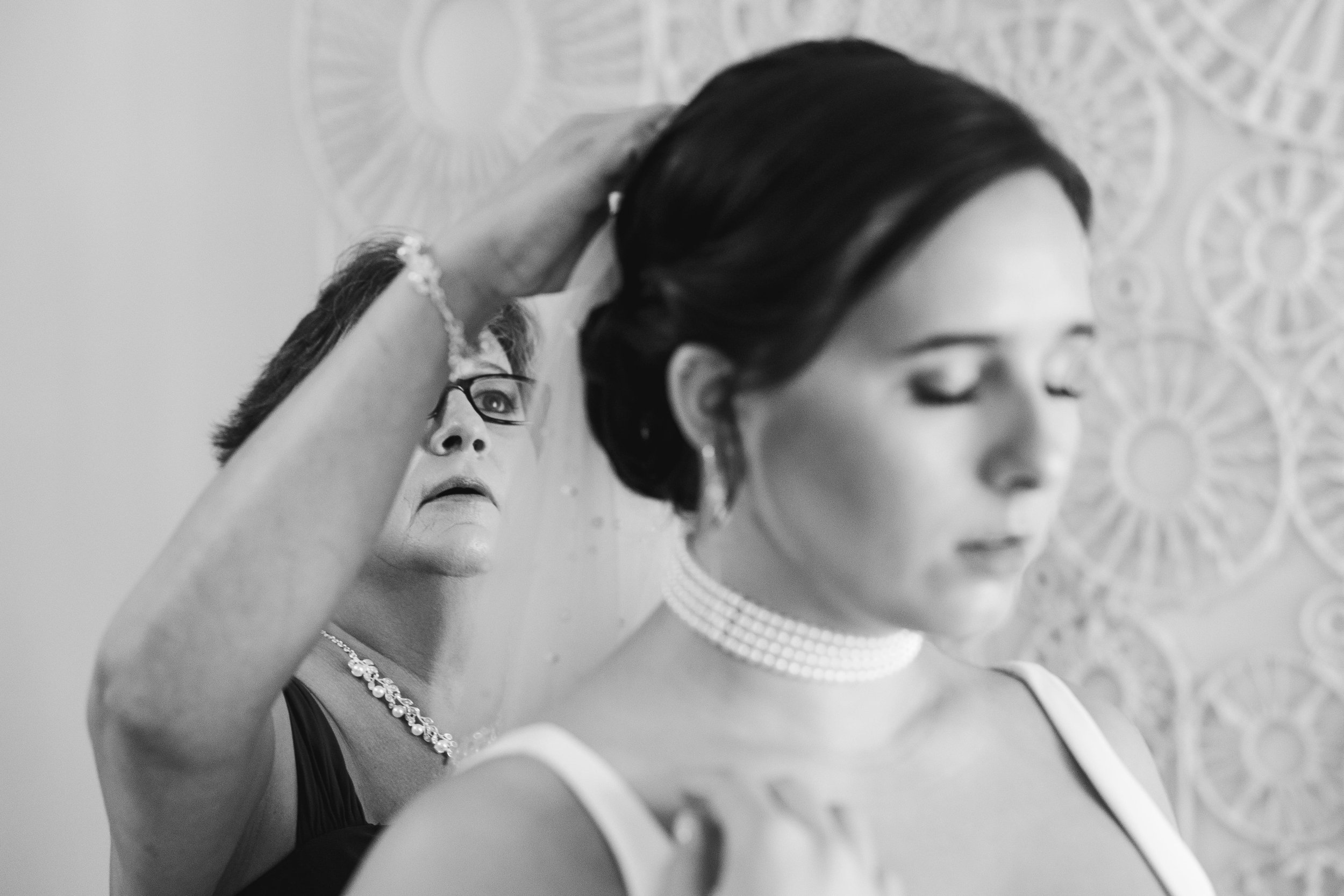 A woman with dark hair wearing a pearl choker and earrings, looking downward, standing next to a mannequin with glasses and jewelry, in black and white.