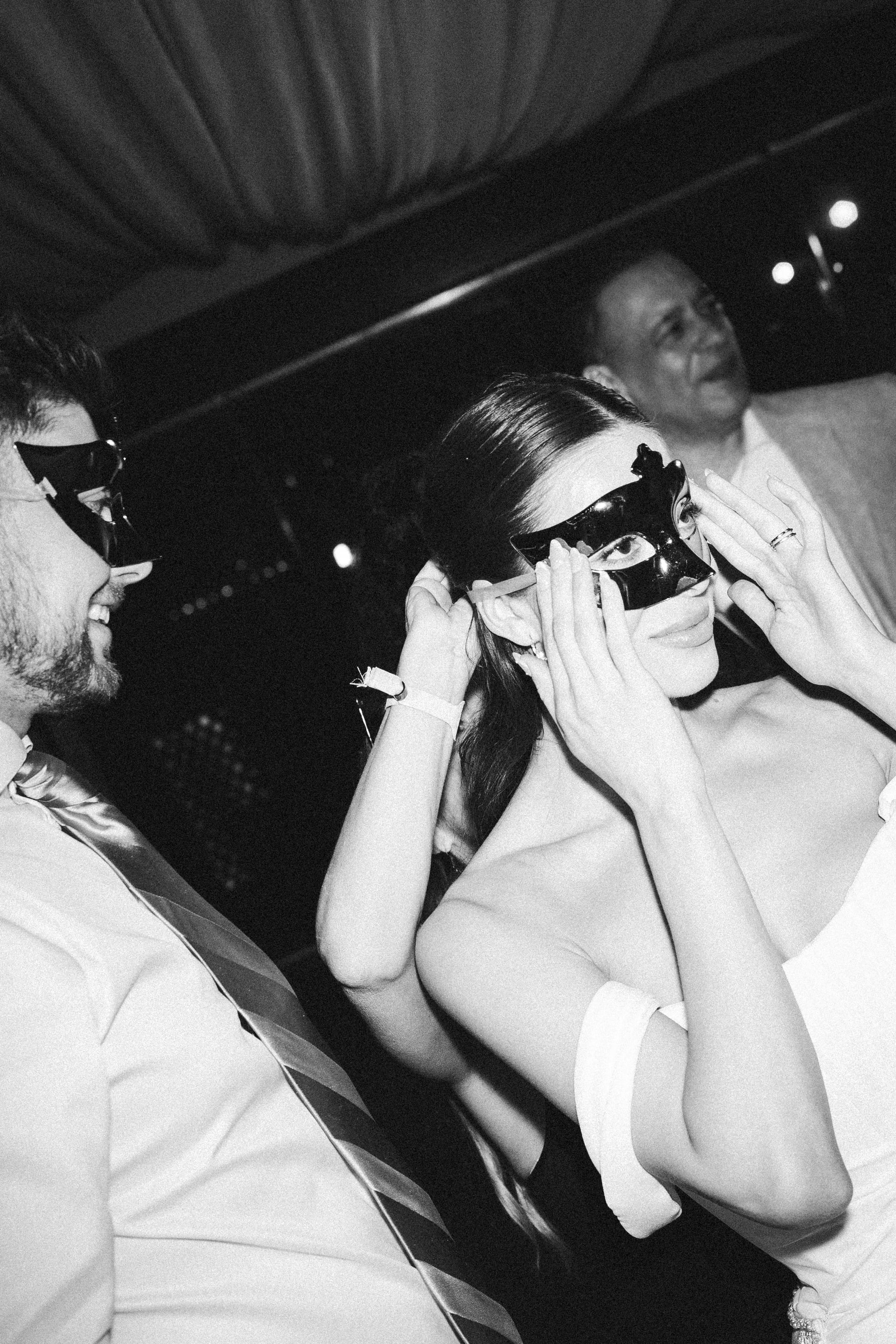 Black and white photo of three people sitting in a vehicle, two women wearing masks and sunglasses, smiling and talking, one man smiling in the background.