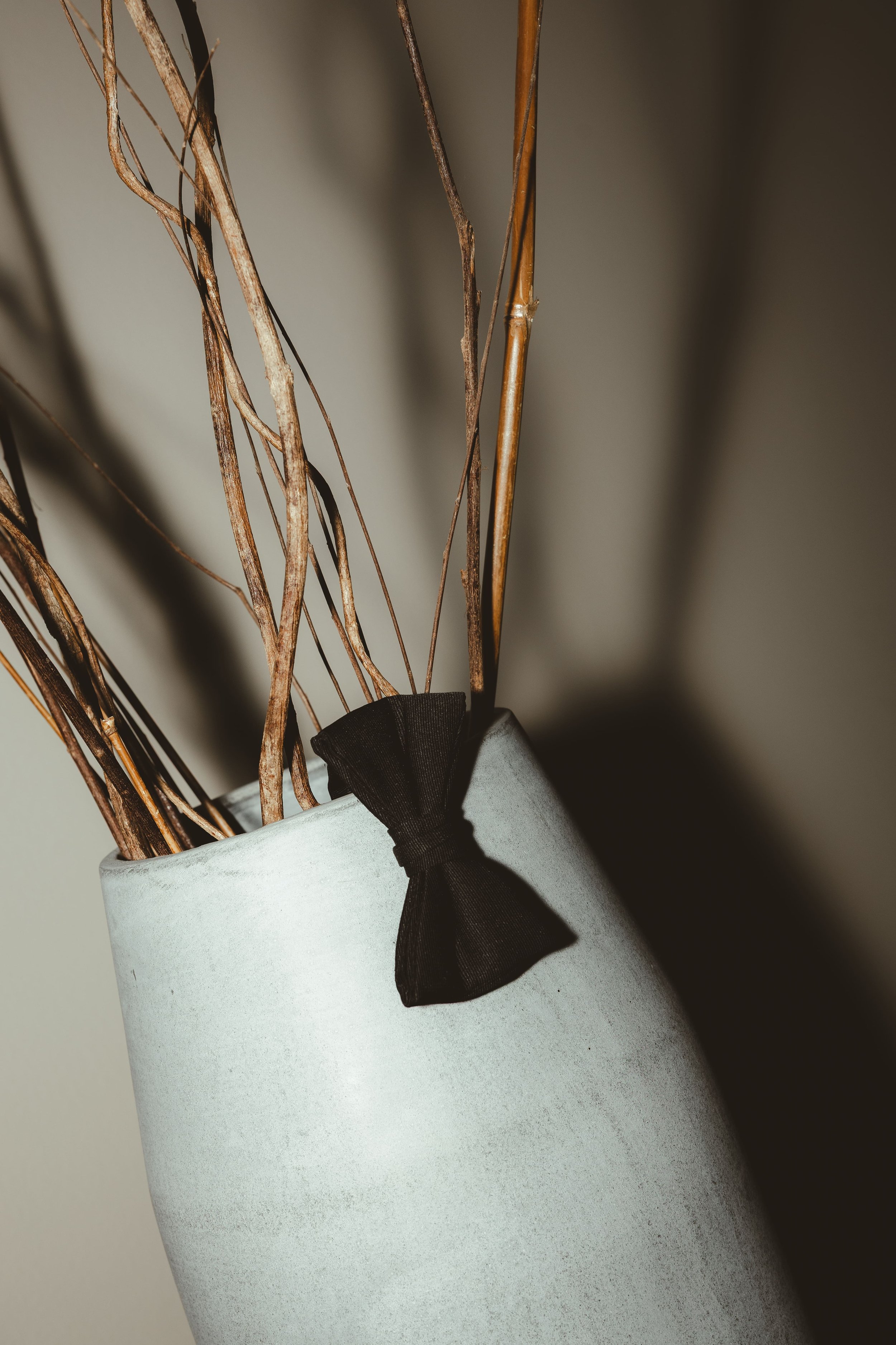 A white ceramic vase with a black ribbon tied around its neck, holding dried brown and beige twigs.
