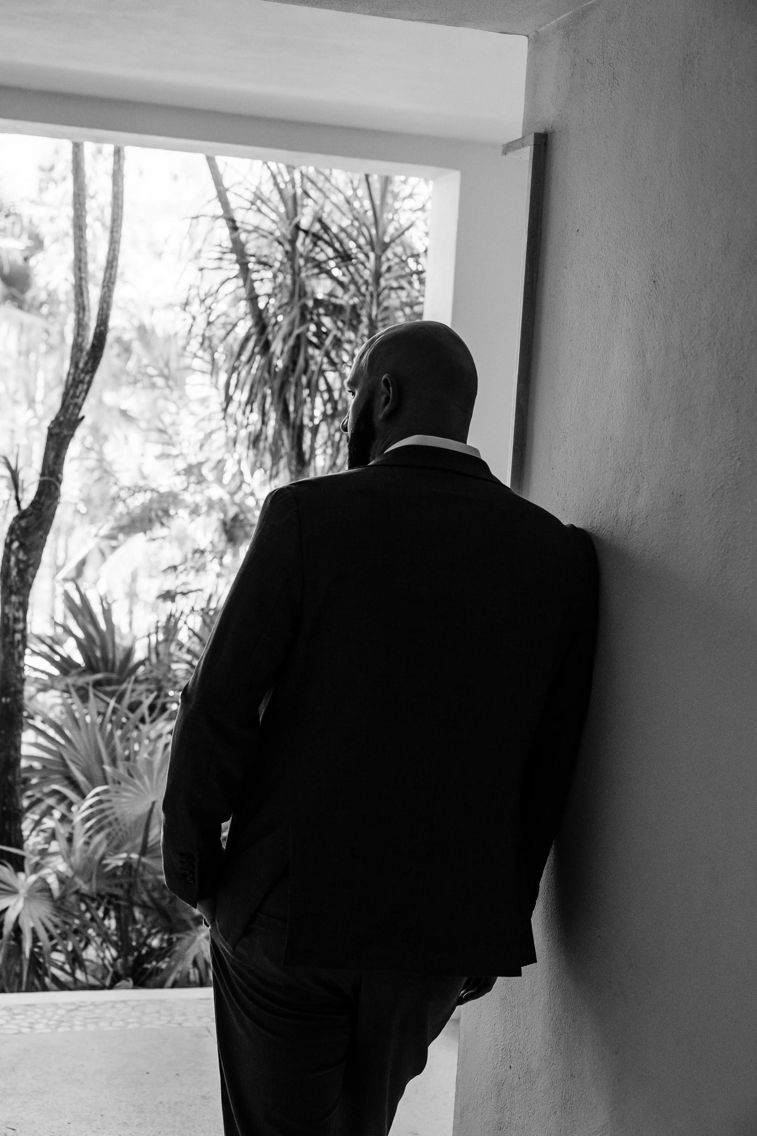 A man in a suit leaning against a wall, seen in profile, with trees and foliage in the background.