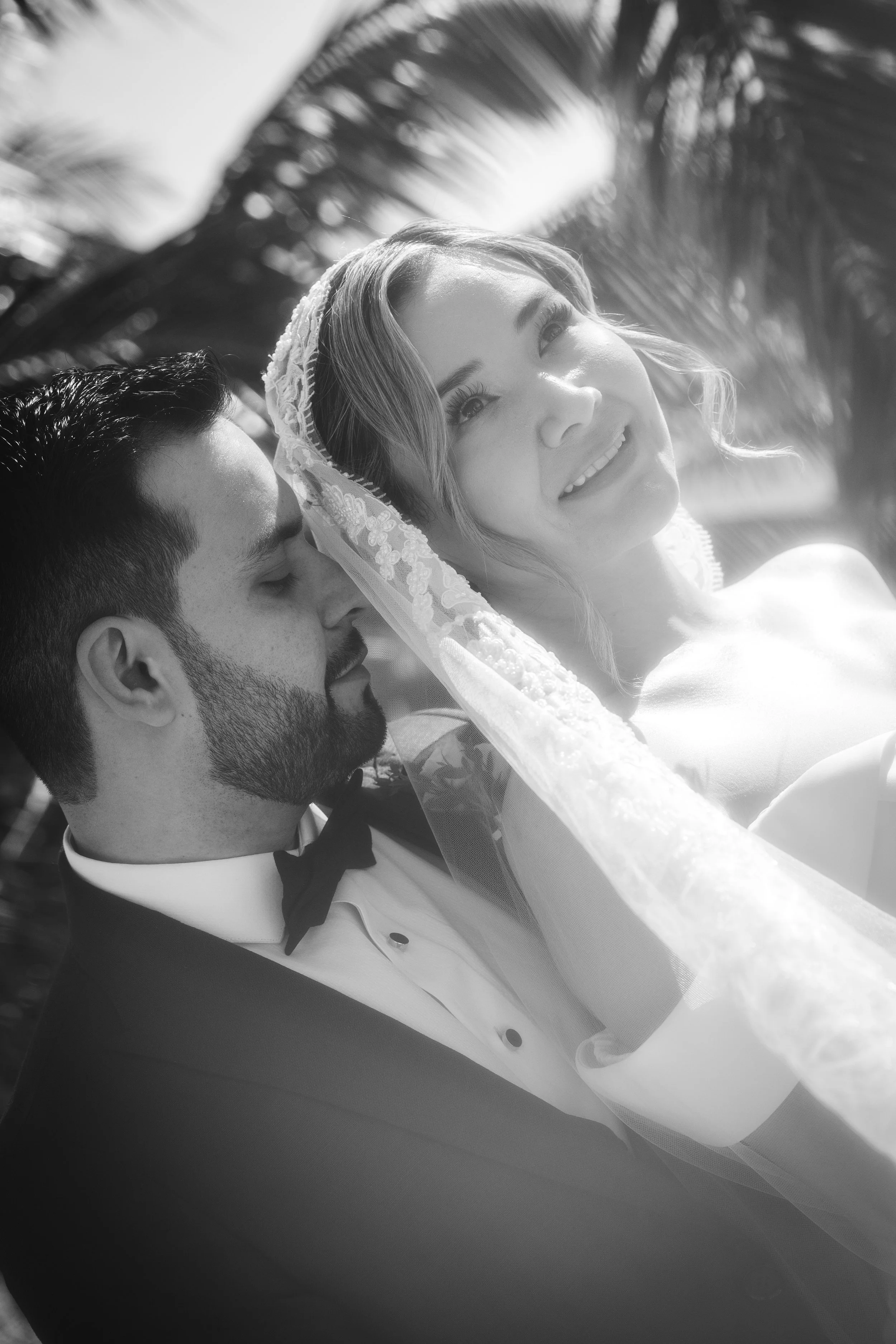 Black and white photo of a bride with a smiling woman, a man with a beard and dark hair, a bow tie, and a tuxedo, set against a background of palm leaves and sunlight filtering through the trees.