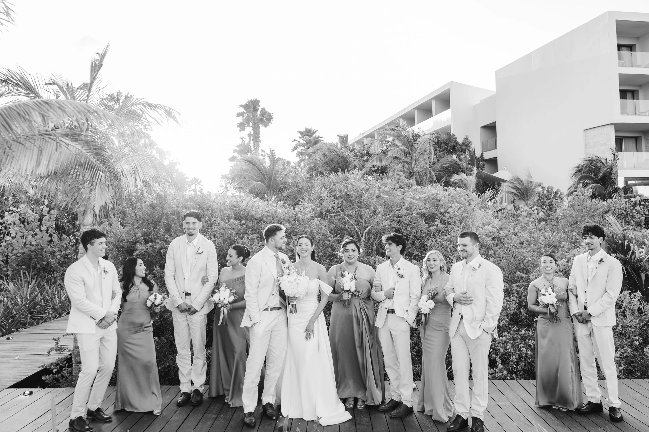 A group of people dressed in formal attire, standing outdoors in front of a lush, tropical landscape with modern white buildings, during daytime.