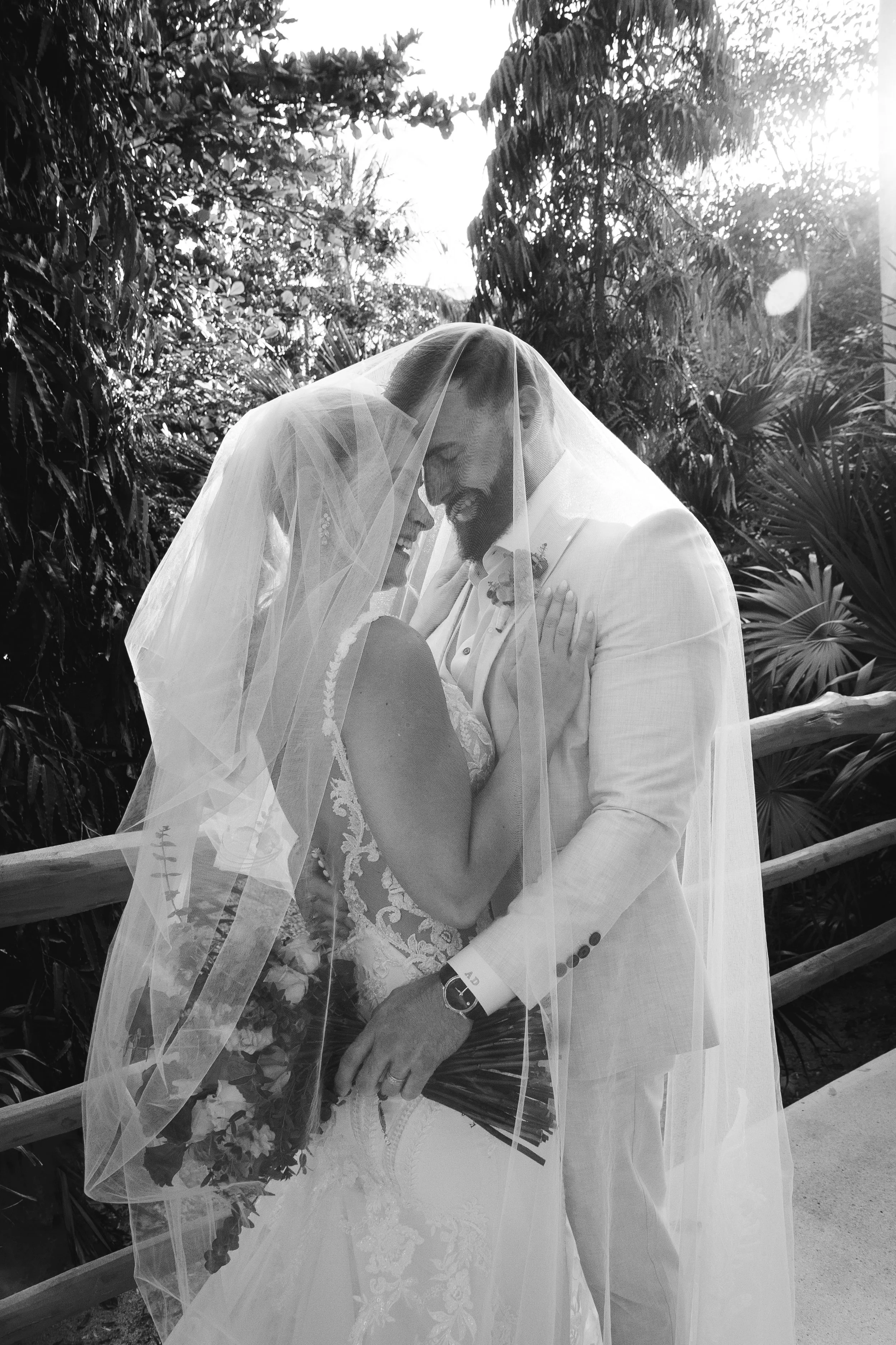 A joyful couple on their wedding day, standing close together under a veil, in an outdoor setting with lush trees and sunlight in the background.