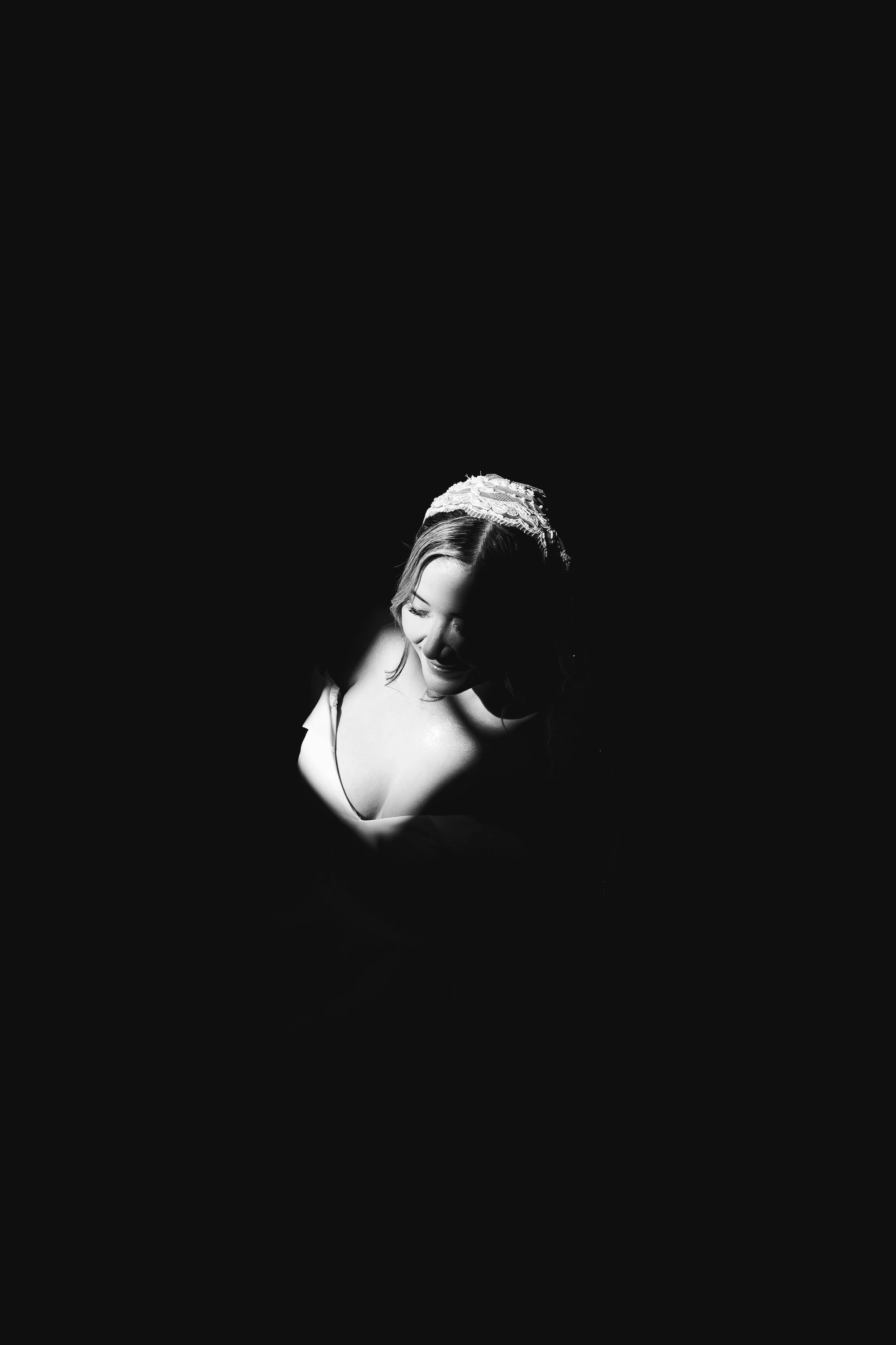 Black and white photo of a smiling woman with dark hair wearing a lacy headband, looking down and illuminated against a dark background.